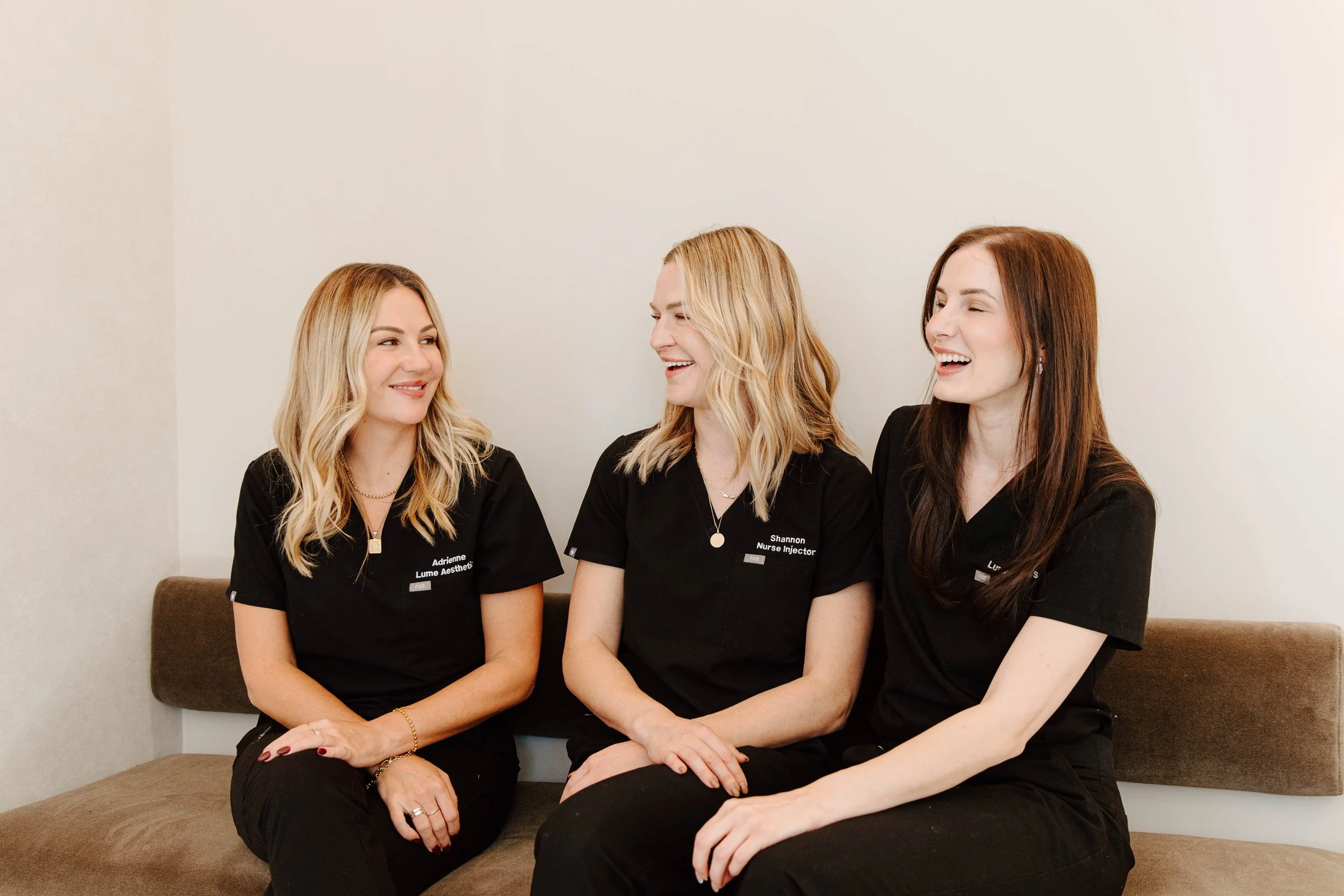 Three women in black uniforms sitting on a brown bench, smiling and engaging in conversation against a plain white wall.