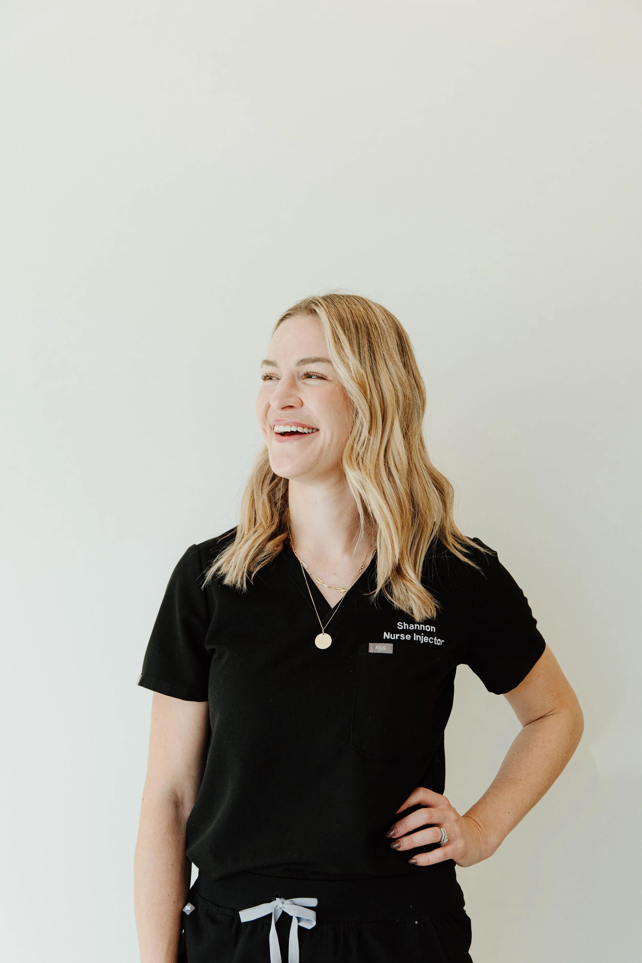 A young woman with blonde hair, smiling and looking to her left, wearing a black medical scrub top with her name badge that reads 'Shannon Nurse Injector' and a pocket, standing against a plain white background.