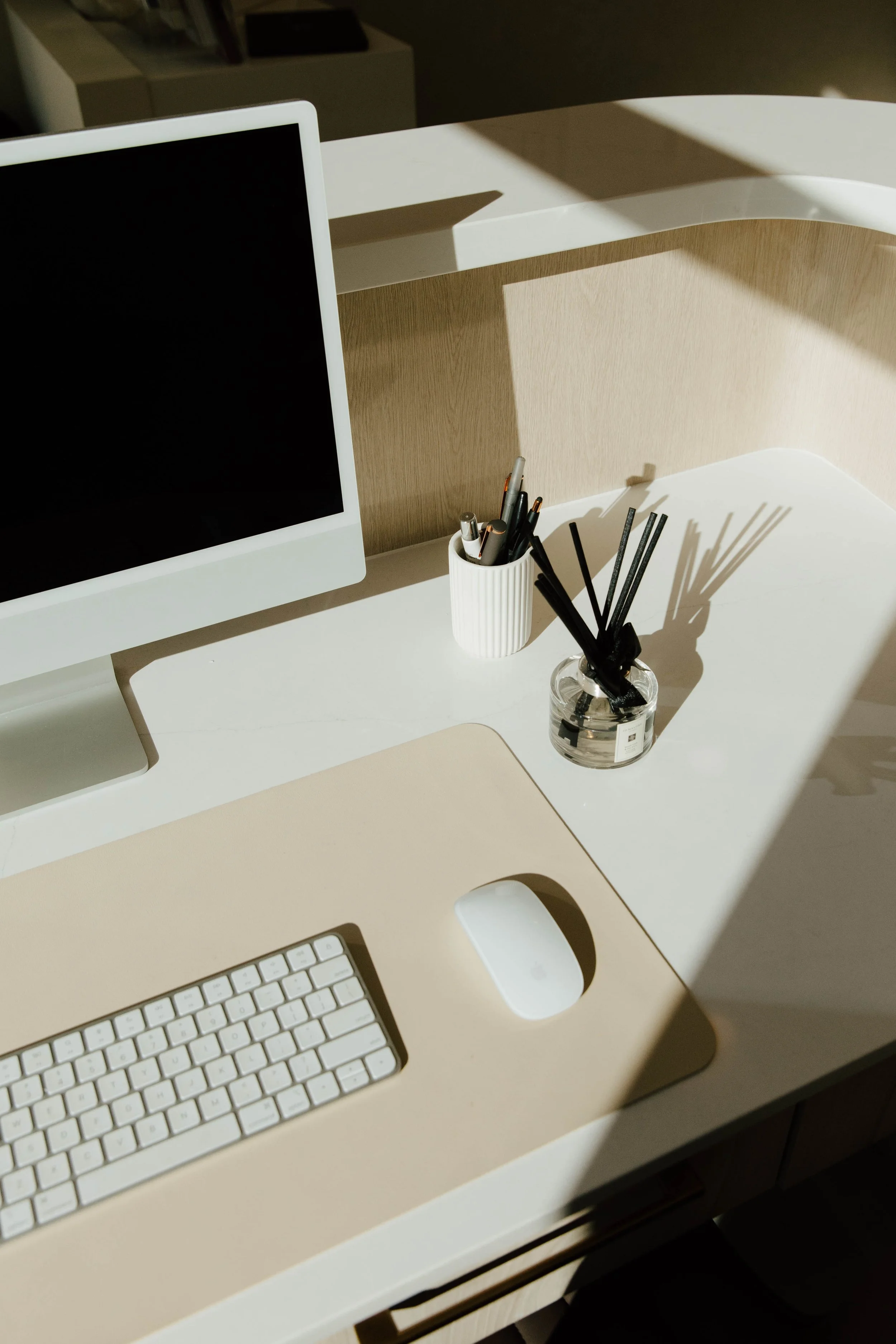 A desktop workspace with a white computer monitor, a beige keyboard, a white mouse on a beige mouse pad, and two containers holding pens and pencils, with sunlight casting shadows across the desk.