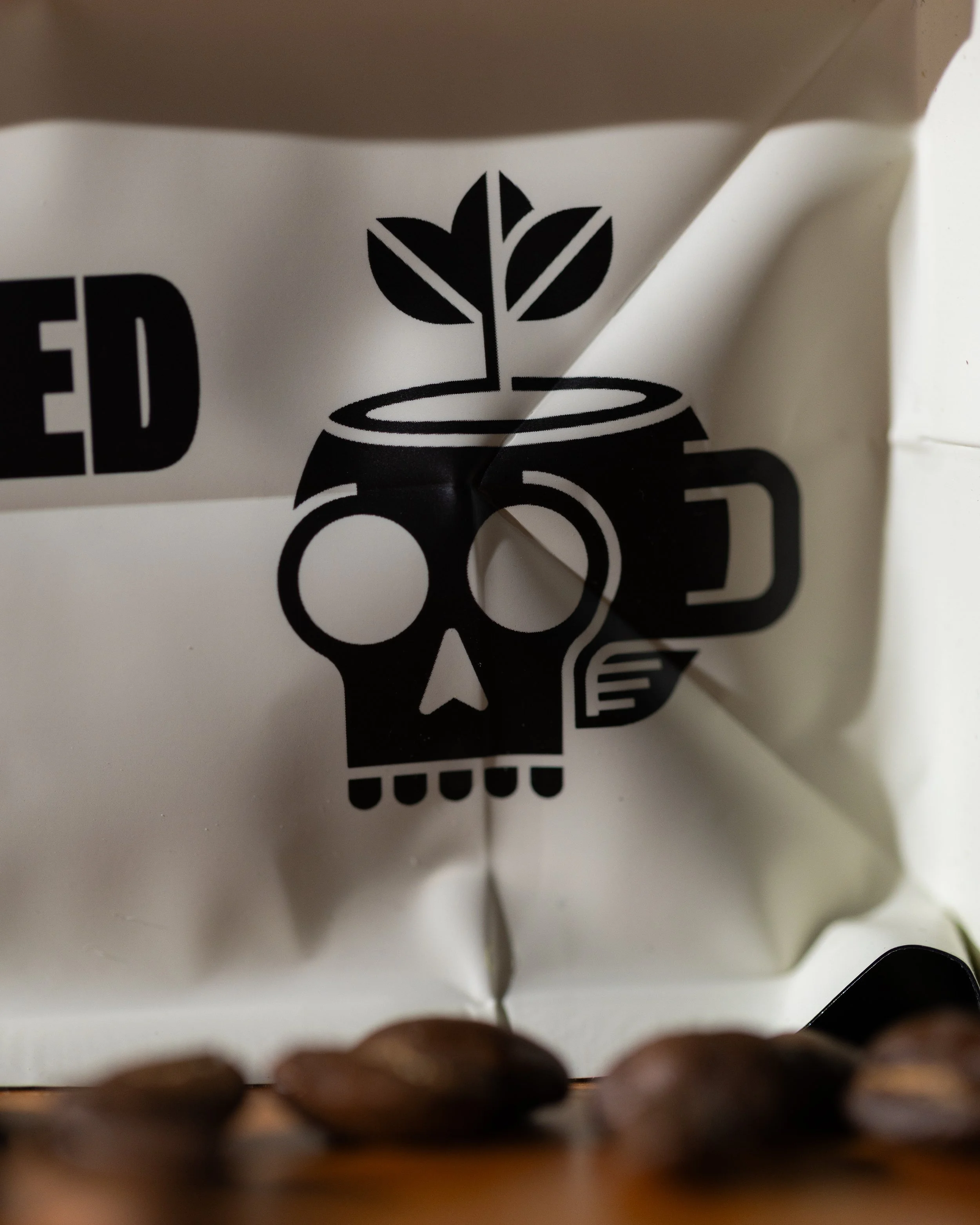 Close-up of a white coffee bag with a black skull and plant design and a part of the word "Brewed" visible on the packaging. Several coffee beans are in front of the bag.