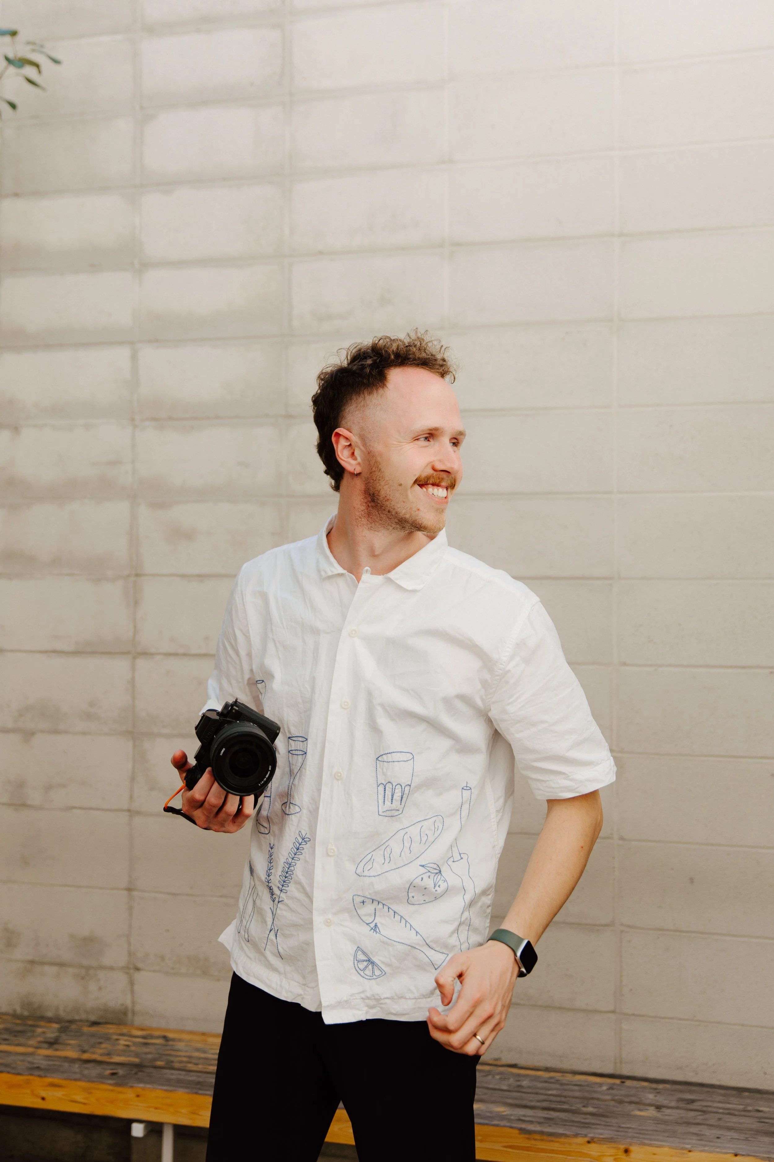 A smiling man with curly hair, wearing a white shirt with blue illustrations of food and drinks, holding a camera in his right hand, standing against a beige brick wall.