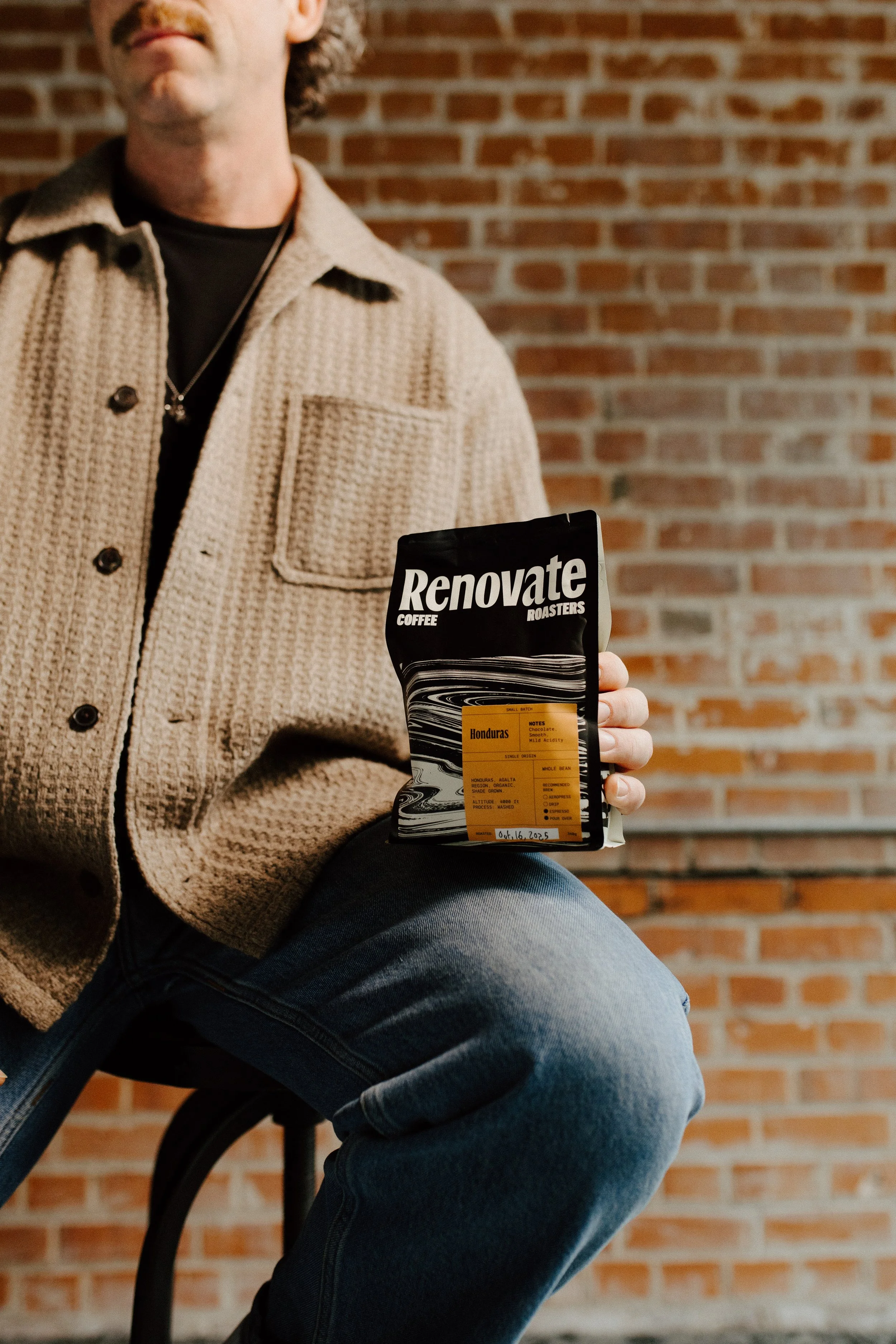 Man in beige textured jacket sitting on chair, holding a bag of coffee labeled 'Renovate Coffee Roasters' in front of a brick wall.