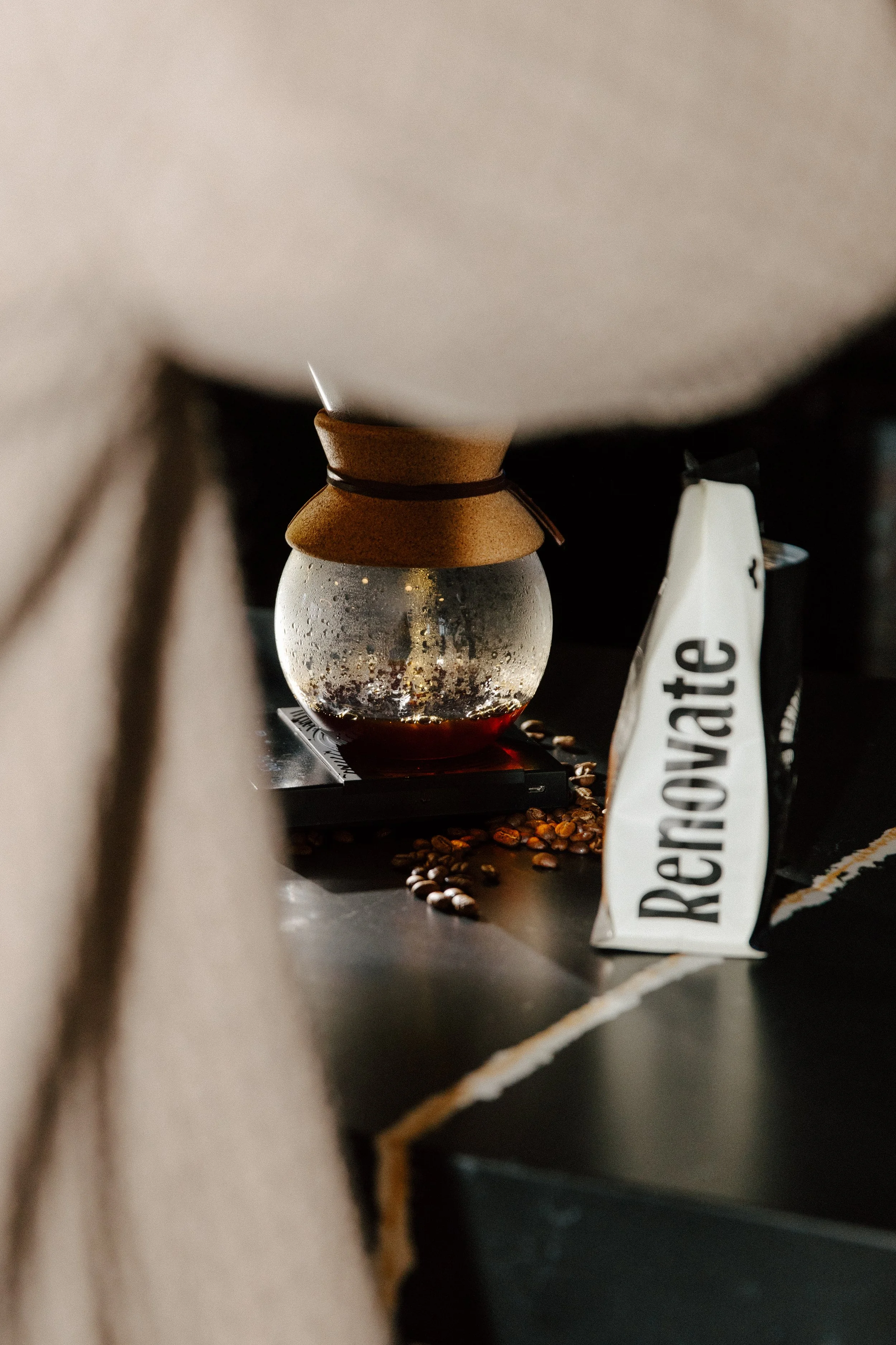 A glass coffee dripper with brewed coffee in it, placed on a scale, with coffee beans scattered around and a bag of Re:Vive coffee next to it.