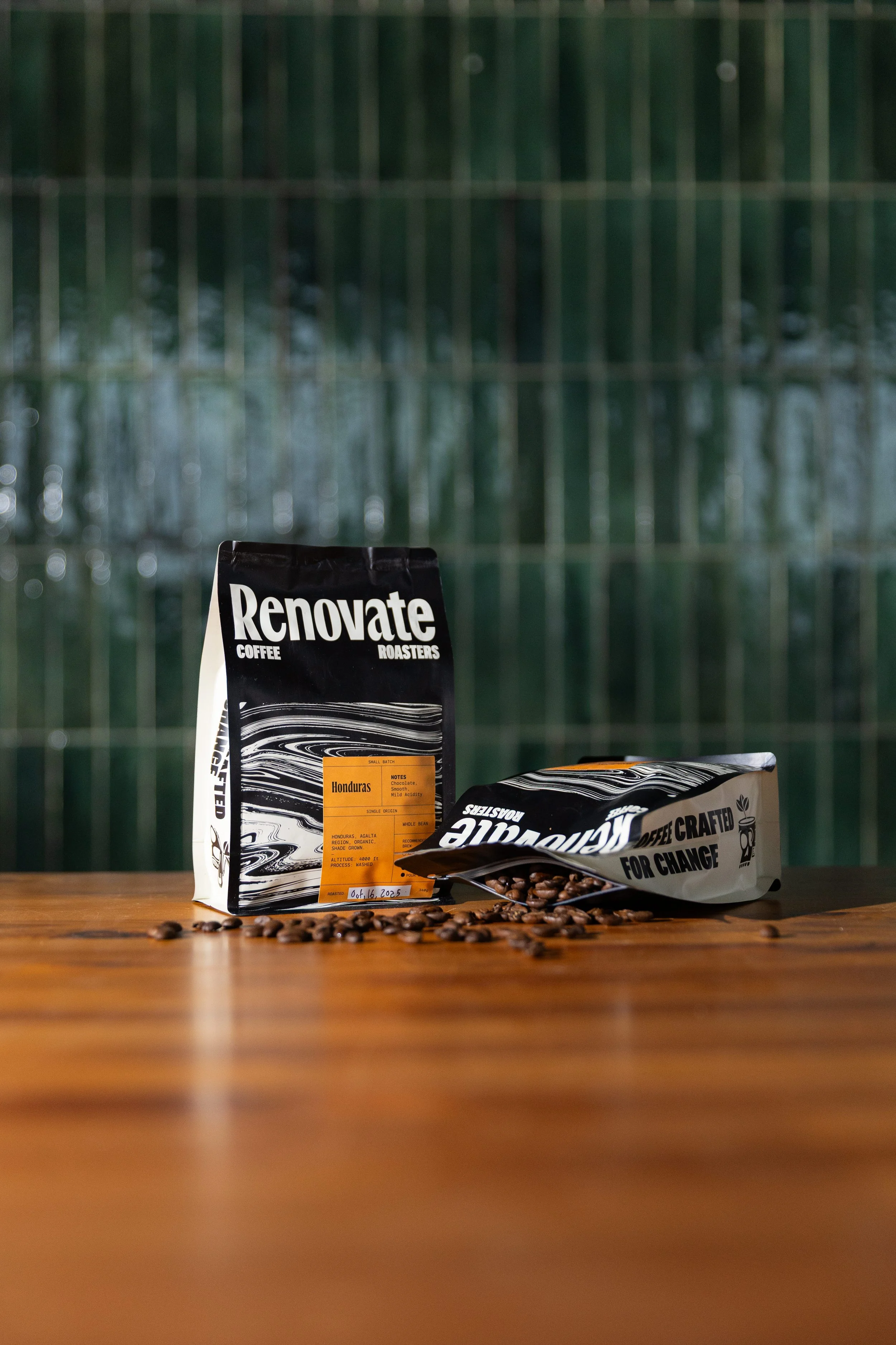 A black and white bag of coffee labeled 'Renovate Coffee Roasters' standing upright and a partially opened bag of coffee beans lying on its side with coffee beans spilled on a wooden surface.