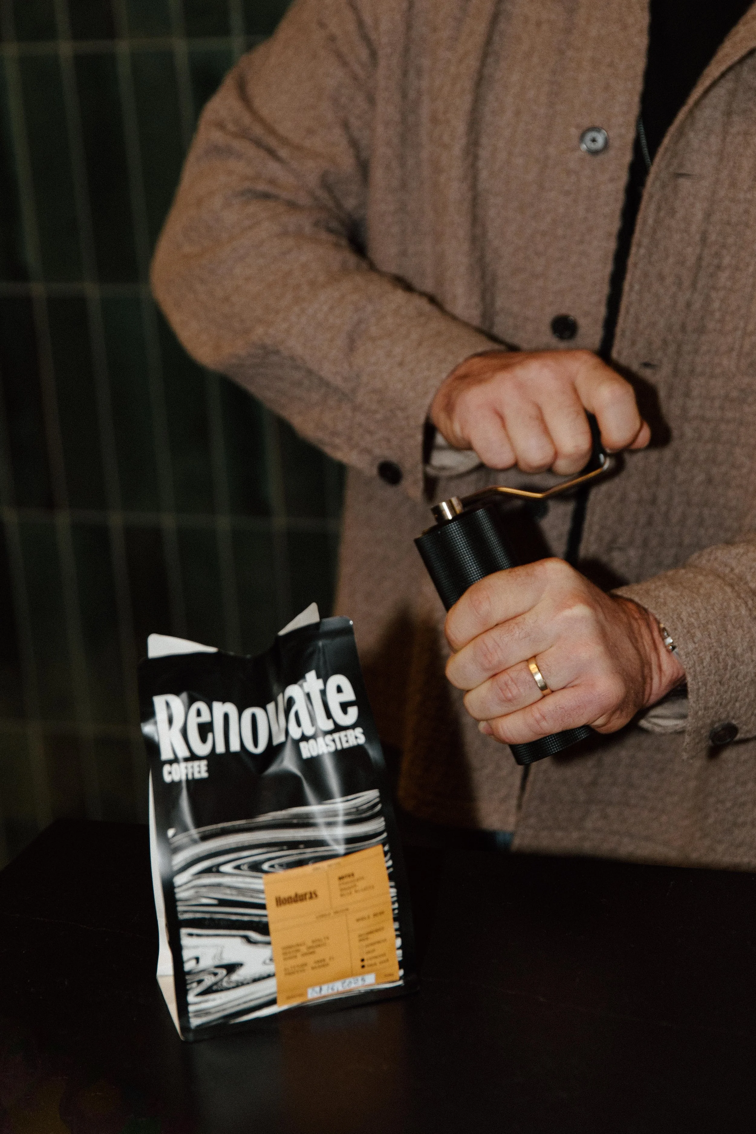 A person in a brown coat is using a coffee grinder, with a bag of Renovate coffee on the table nearby.