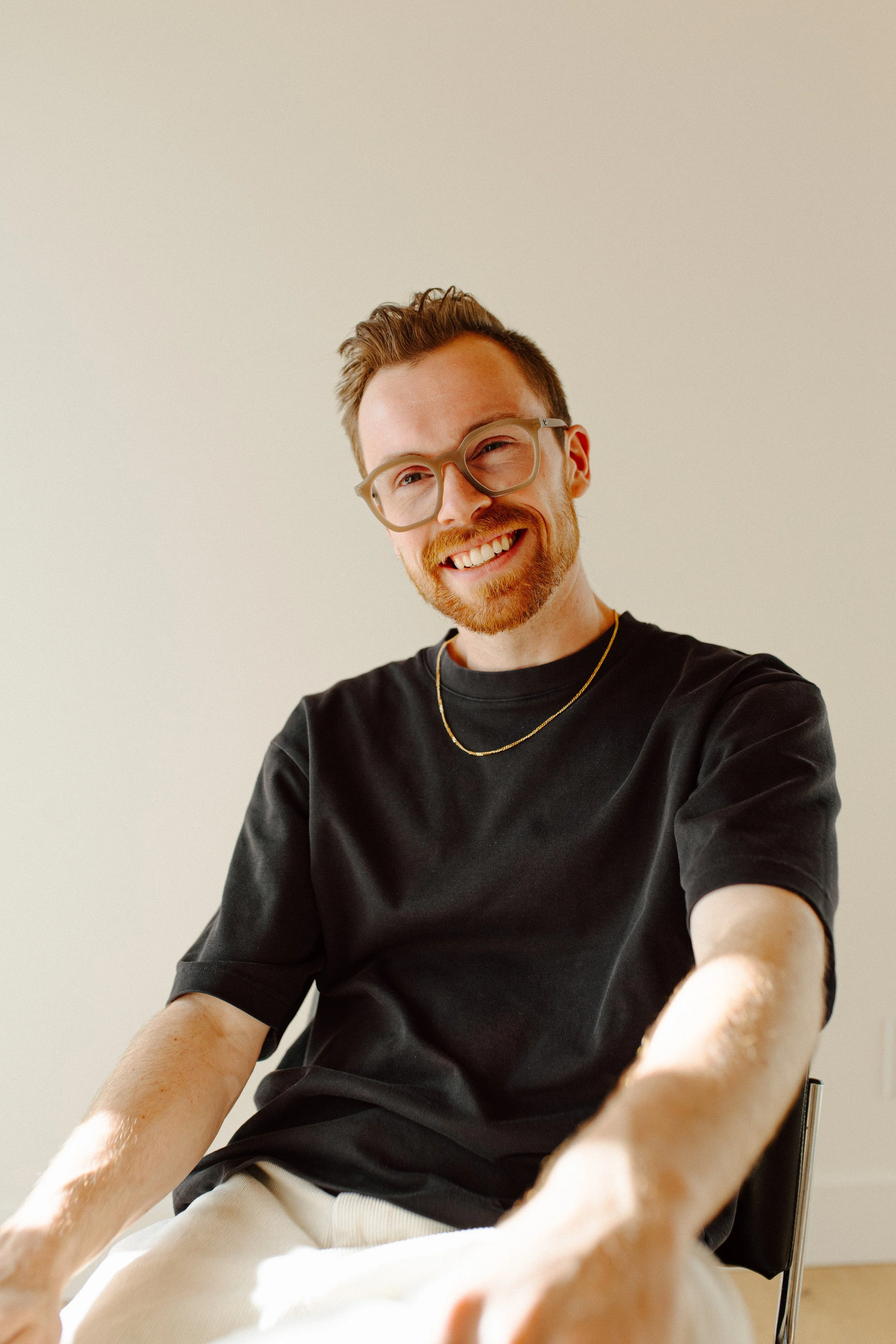 A smiling man with glasses, a beard, wearing a black shirt, a gold chain, and light-colored pants, sitting indoors against a plain wall.