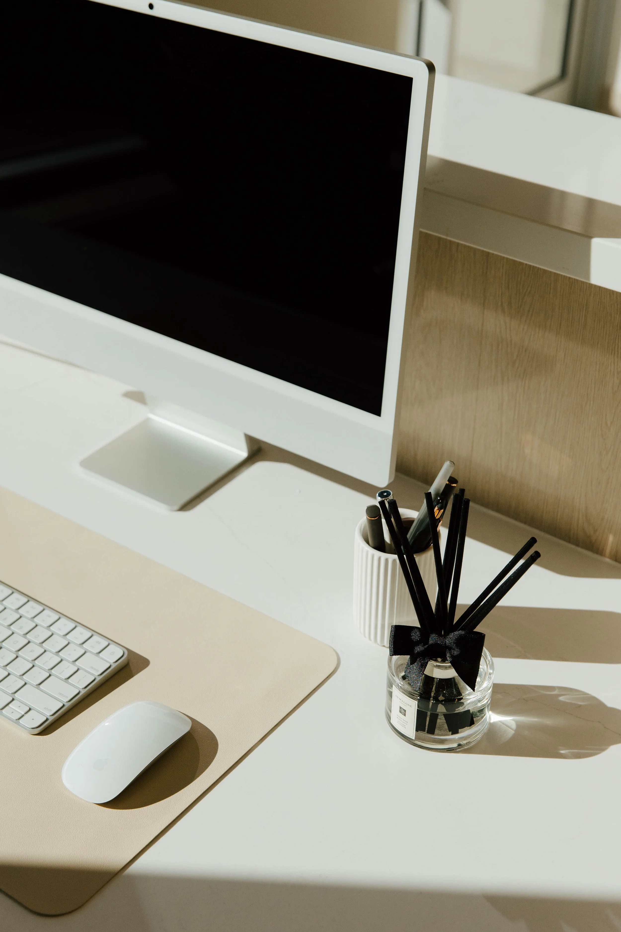 A minimalist workspace with a computer monitor, keyboard, mouse, and small container holding black pens and a white pen, with sunlight casting shadows.