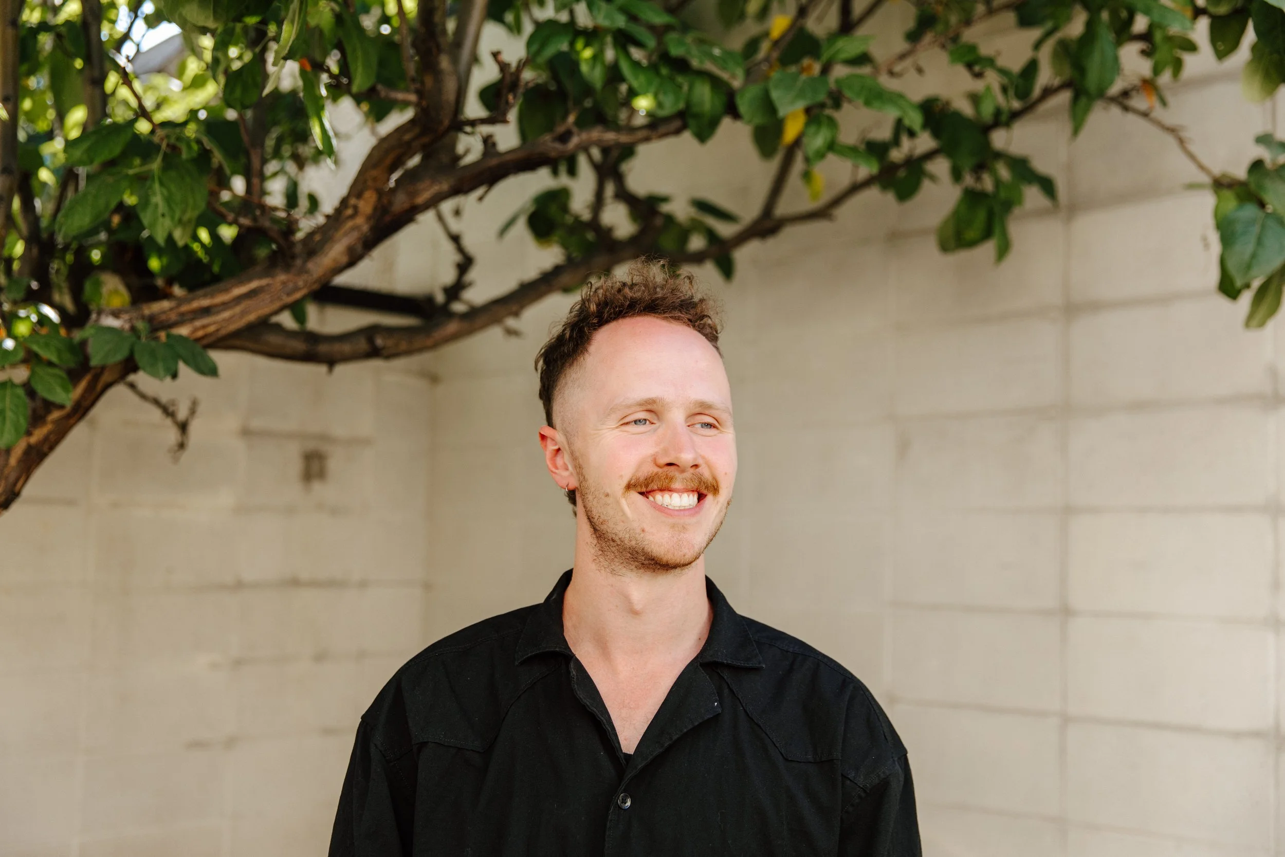 A smiling man with short, curly hair and a beard, wearing a black shirt, standing outdoors near a tree with green leaves and a beige brick wall in the background.