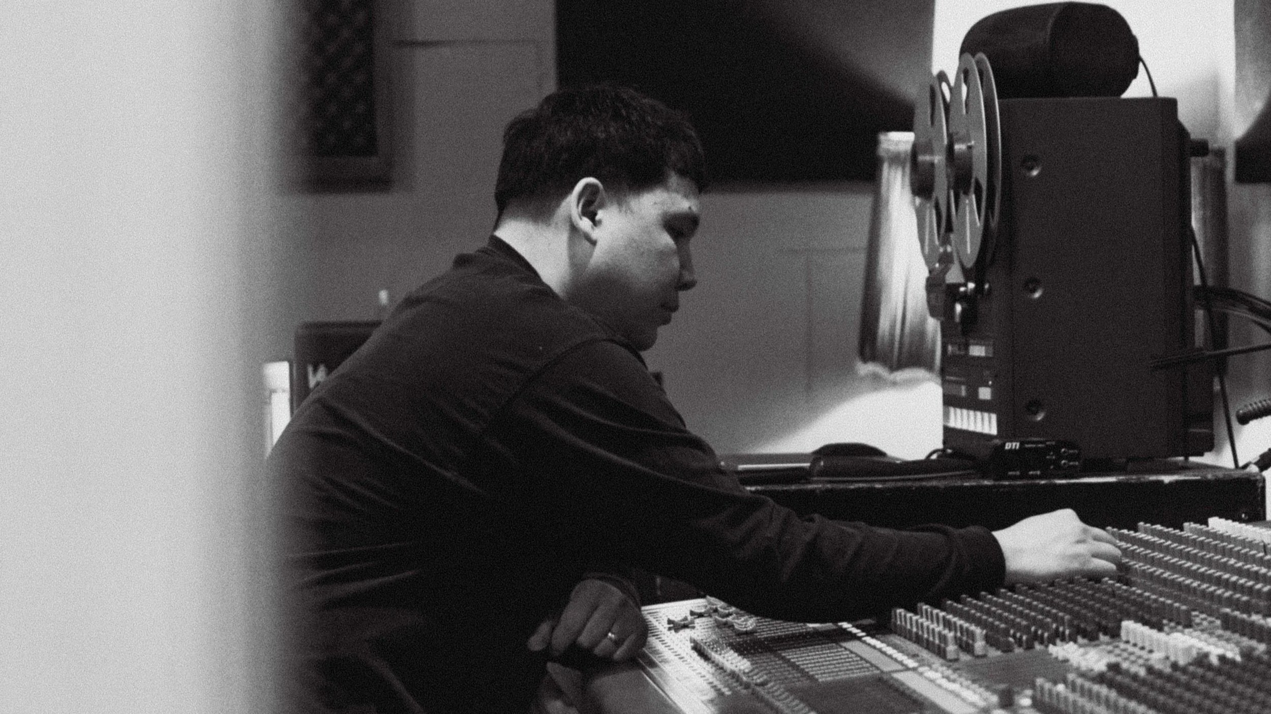Black and white photo of a person focusing on a music mixing console, with a reel-to-reel tape recorder visible on a table.