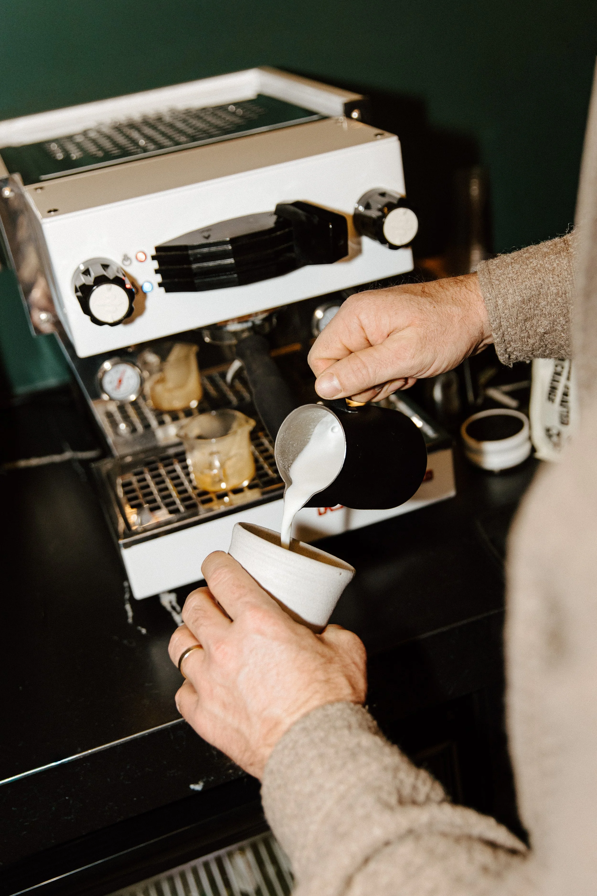 Person pouring steamed milk into a coffee cup from a black pitcher, with a commercial espresso machine in the background.