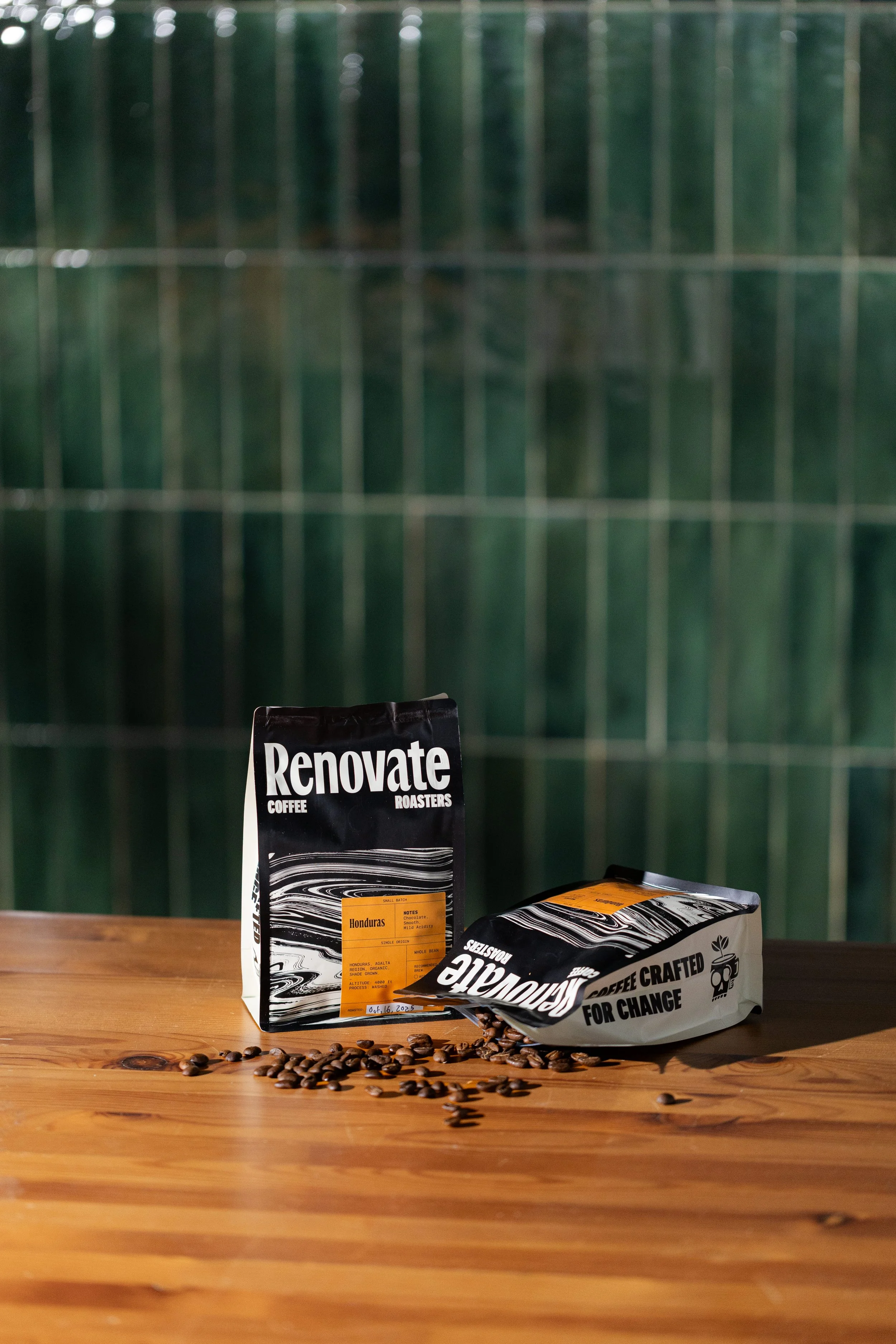 Two bags of coffee beans on a wooden table, with some scattered coffee beans in front, positioned against a green tiled wall background.