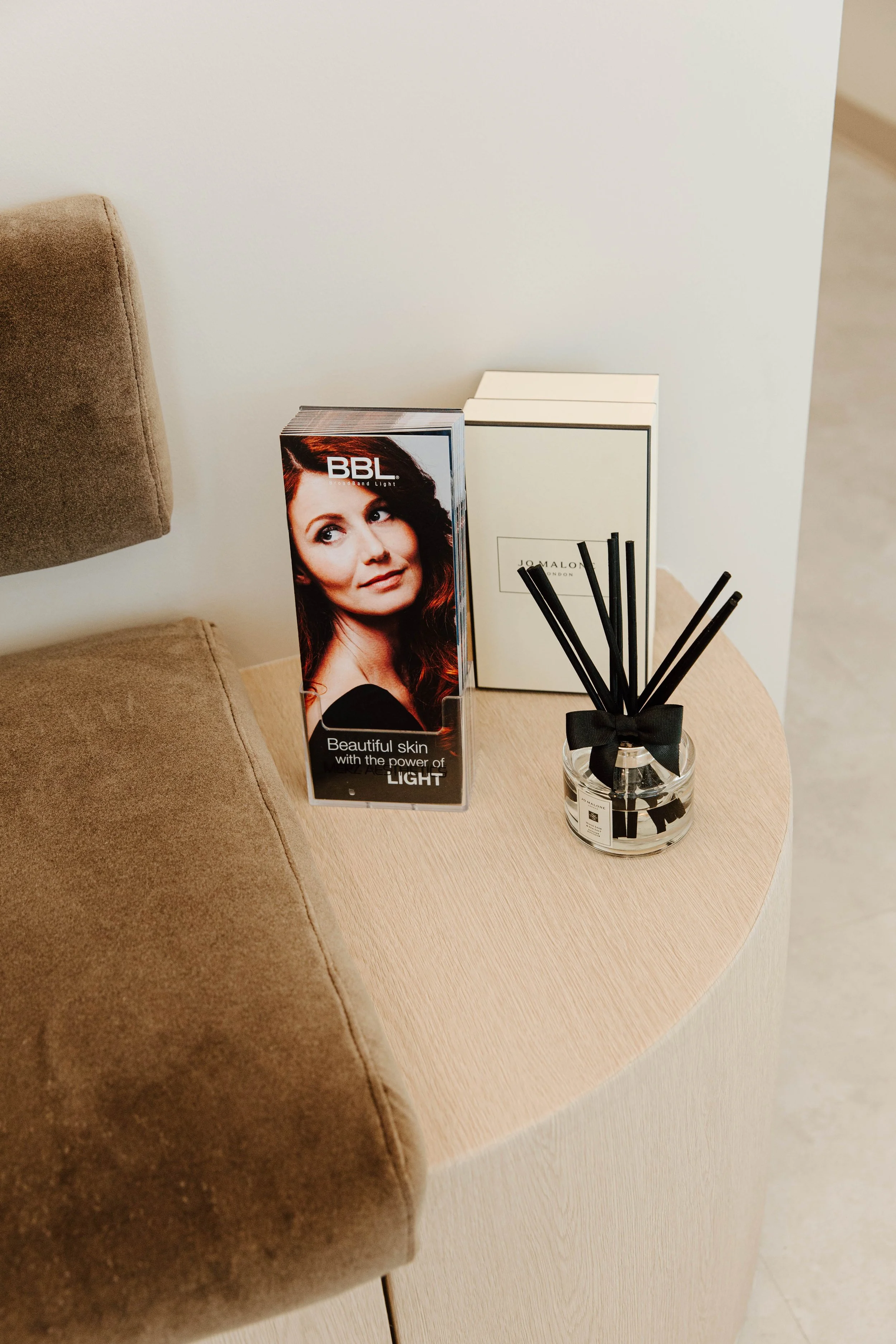 A round wooden side table next to a brown upholstered chair with promotional material, a white box, and a glass container with black reed diffuser sticks and a black ribbon.
