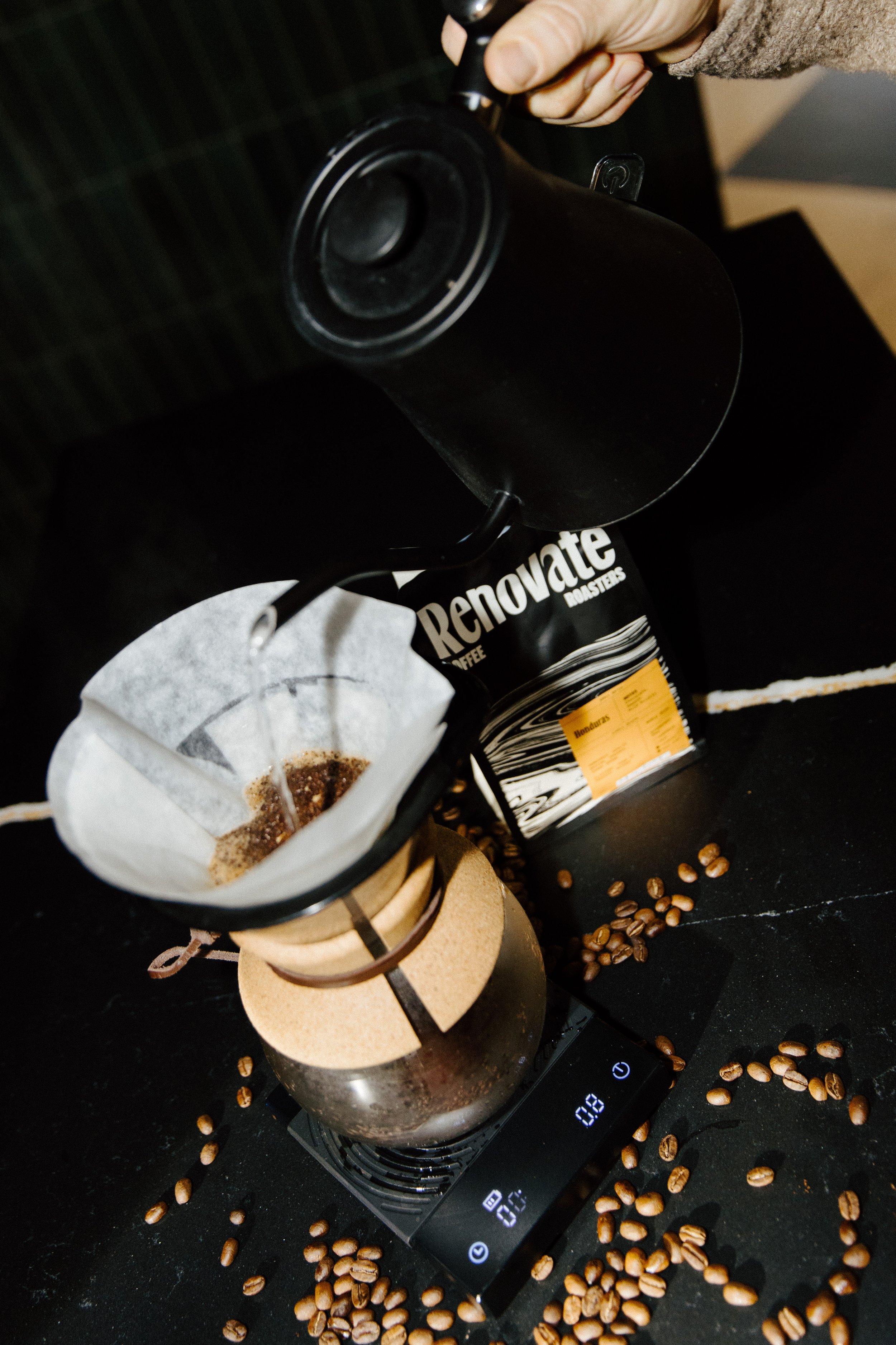 A pour-over coffee maker brewing coffee with a paper filter, a glass carafe, and a bag of Renavate roast coffee beans on a digital scale, with coffee beans scattered around.