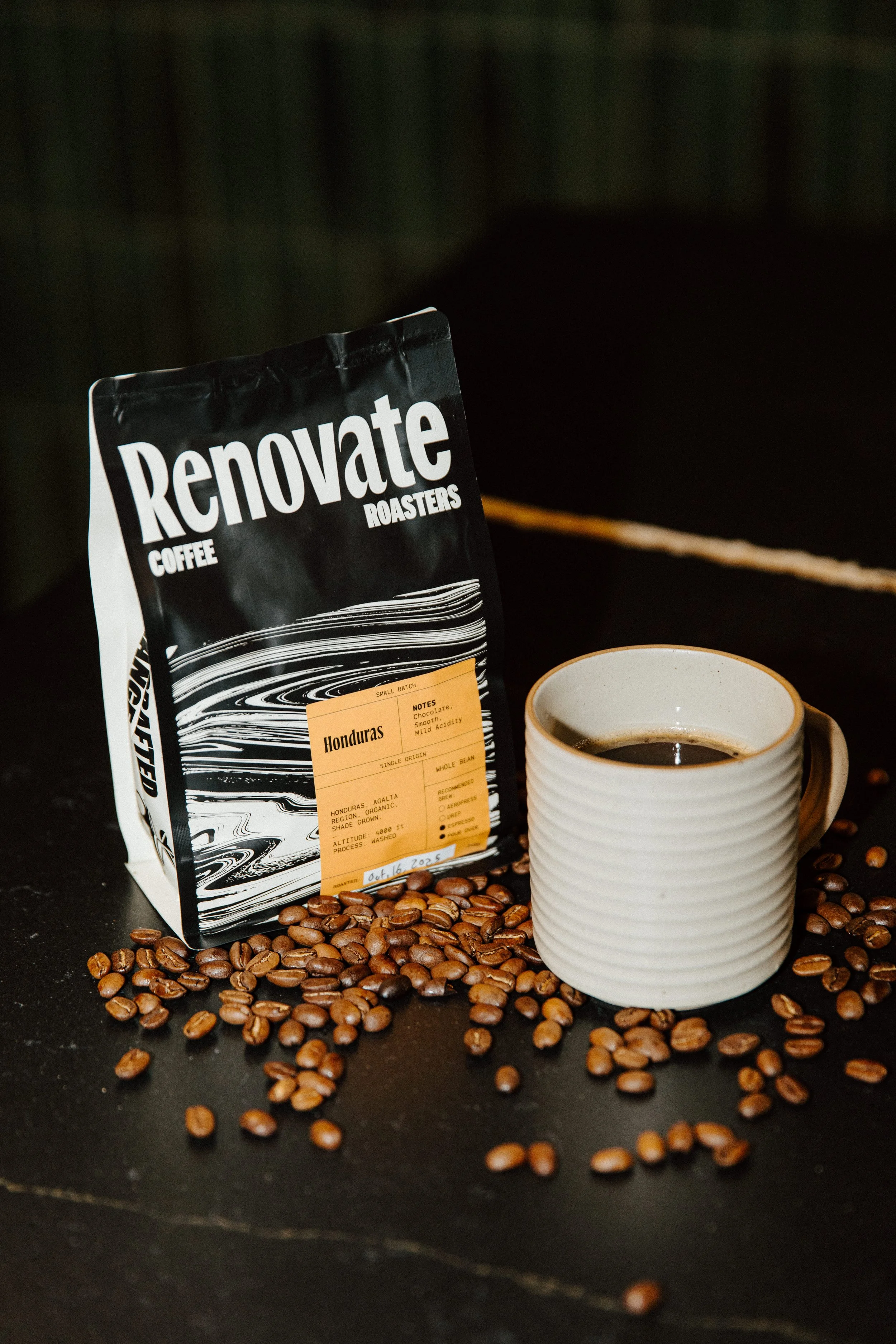 A bag of Renovate Coffee Roasters Honduras coffee beans next to a mug of brewed coffee and scattered coffee beans on a dark surface.