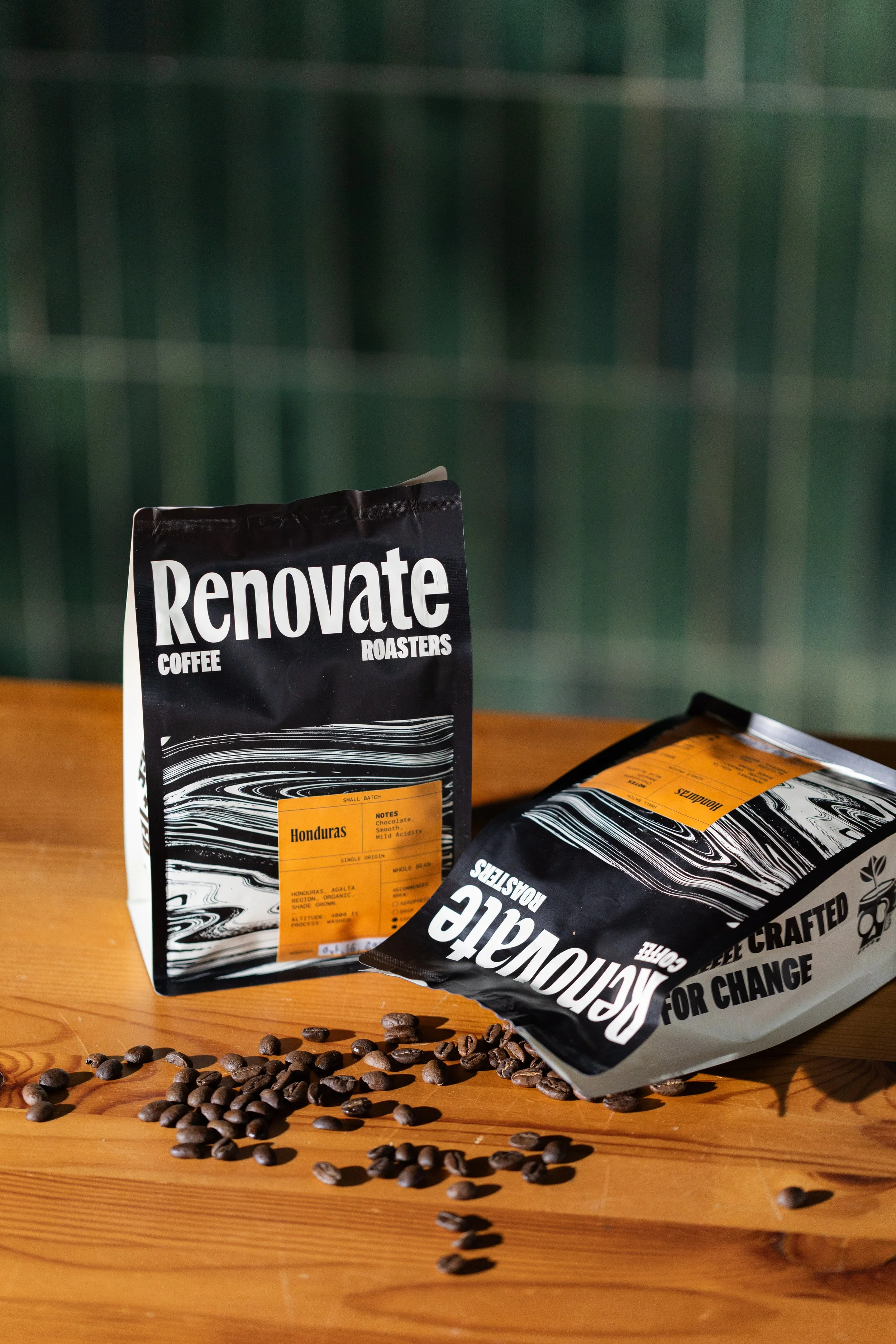 Two bags of RenovatE coffee on a wooden surface with scattered coffee beans, one standing upright and the other lying down, against a green tiled background.