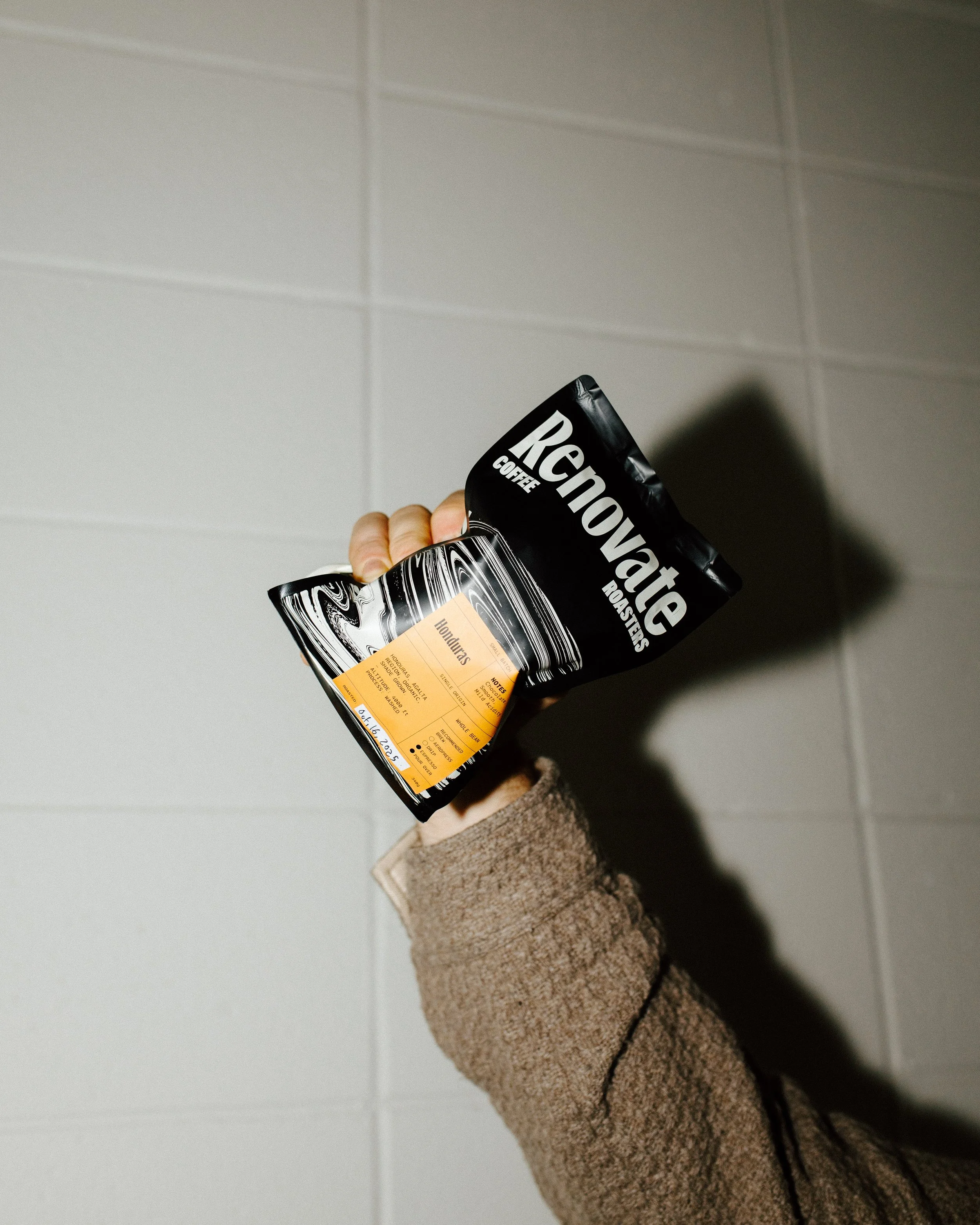 A person holding a black bag of Renevoate Coffee Roasters coffee with a Honduras coffee label against a tiled wall.