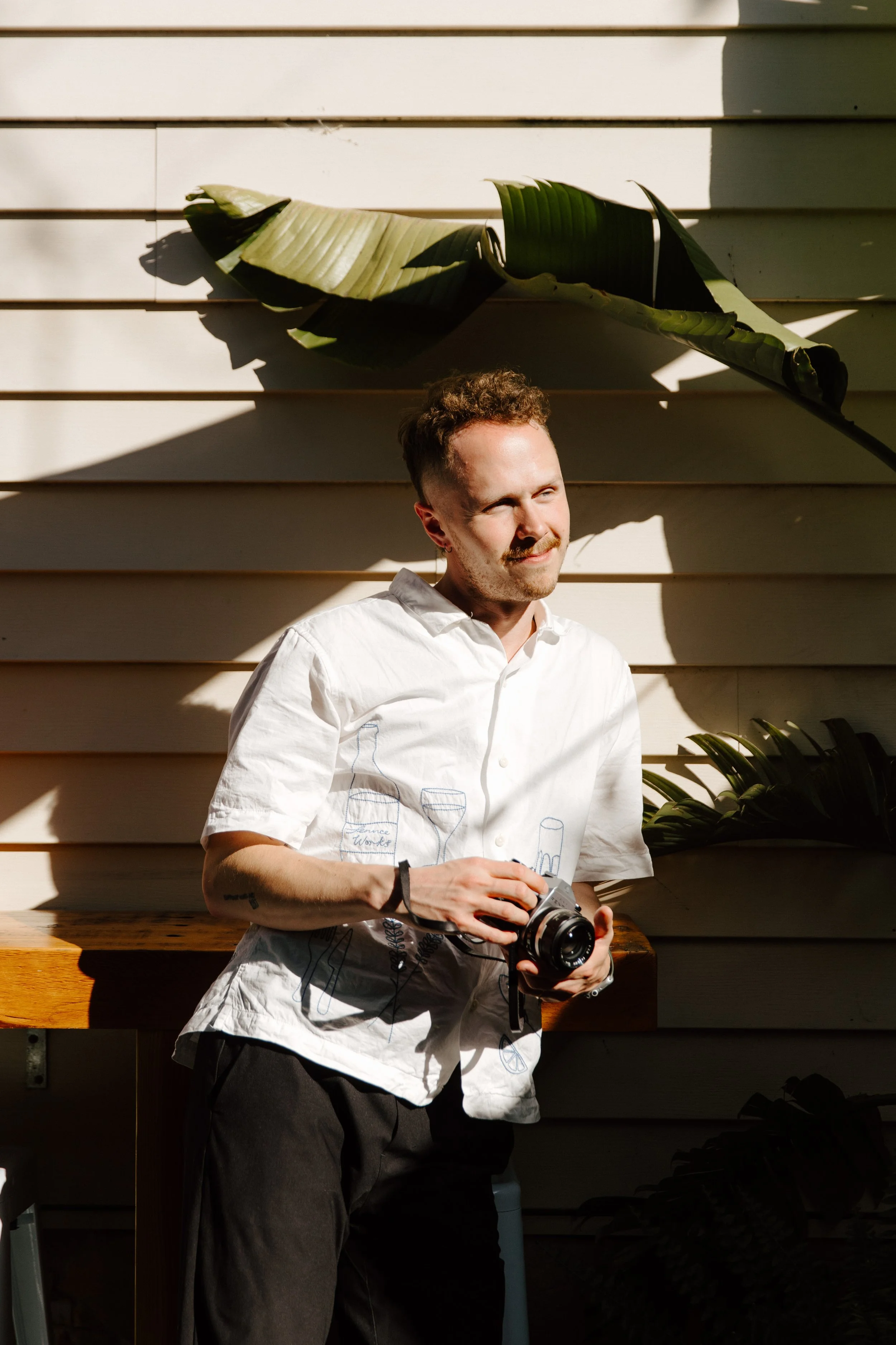 A man holding a camera, standing outdoors against a light-colored wooden wall with shadows and a large green leaf.