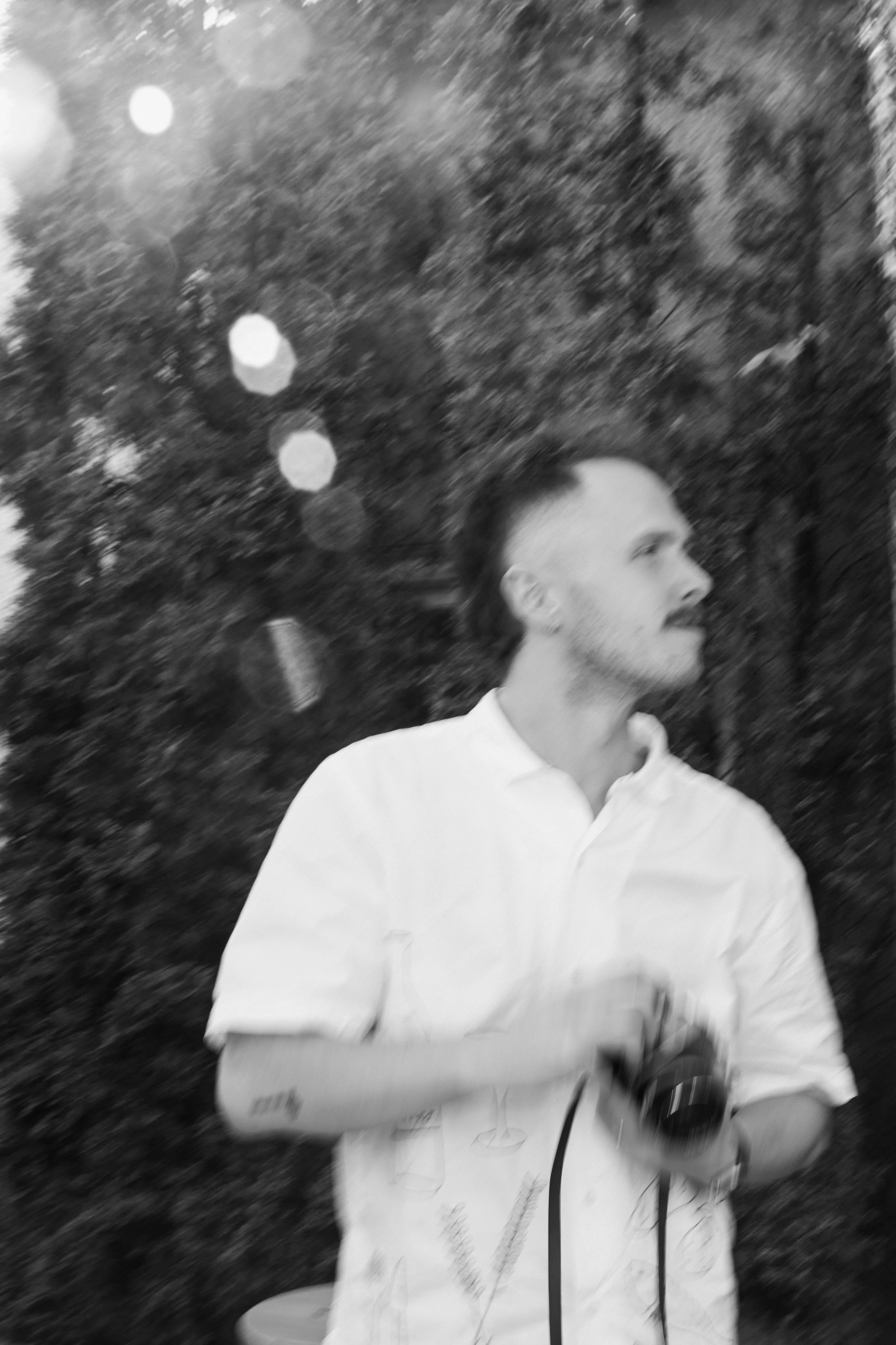 A young man with styled hair and a beard, wearing a white button-up shirt, is standing outdoors holding a camera with his arms crossed. There are blurred trees and bokeh lights in the background.
