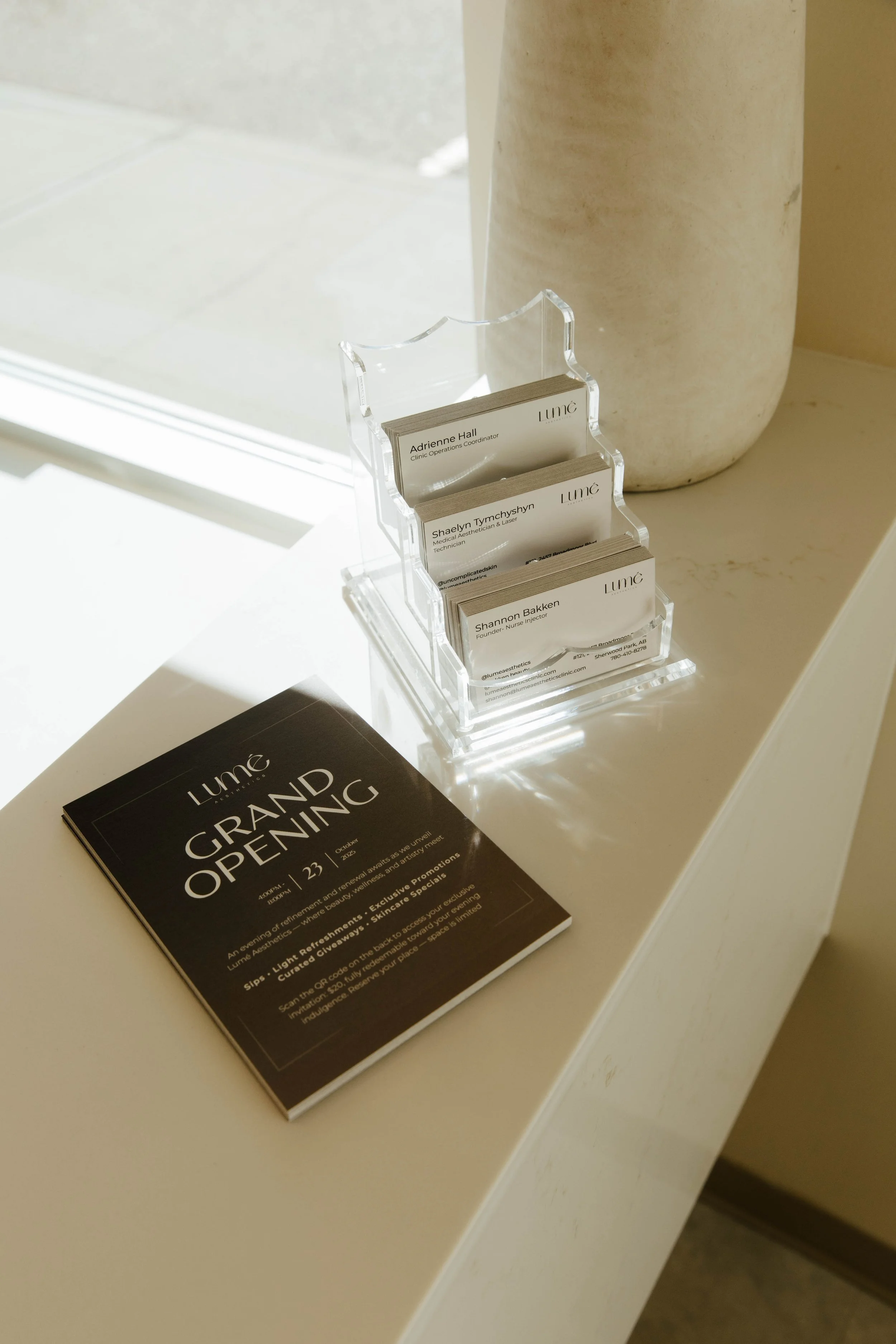 A black flyer with white text on a white countertop reads 'LUME GRAND OPENING'. Next to it, a clear acrylic business card holder contains several business cards for LUME, a medical aesthetic and laser clinic, with some visible names and titles.