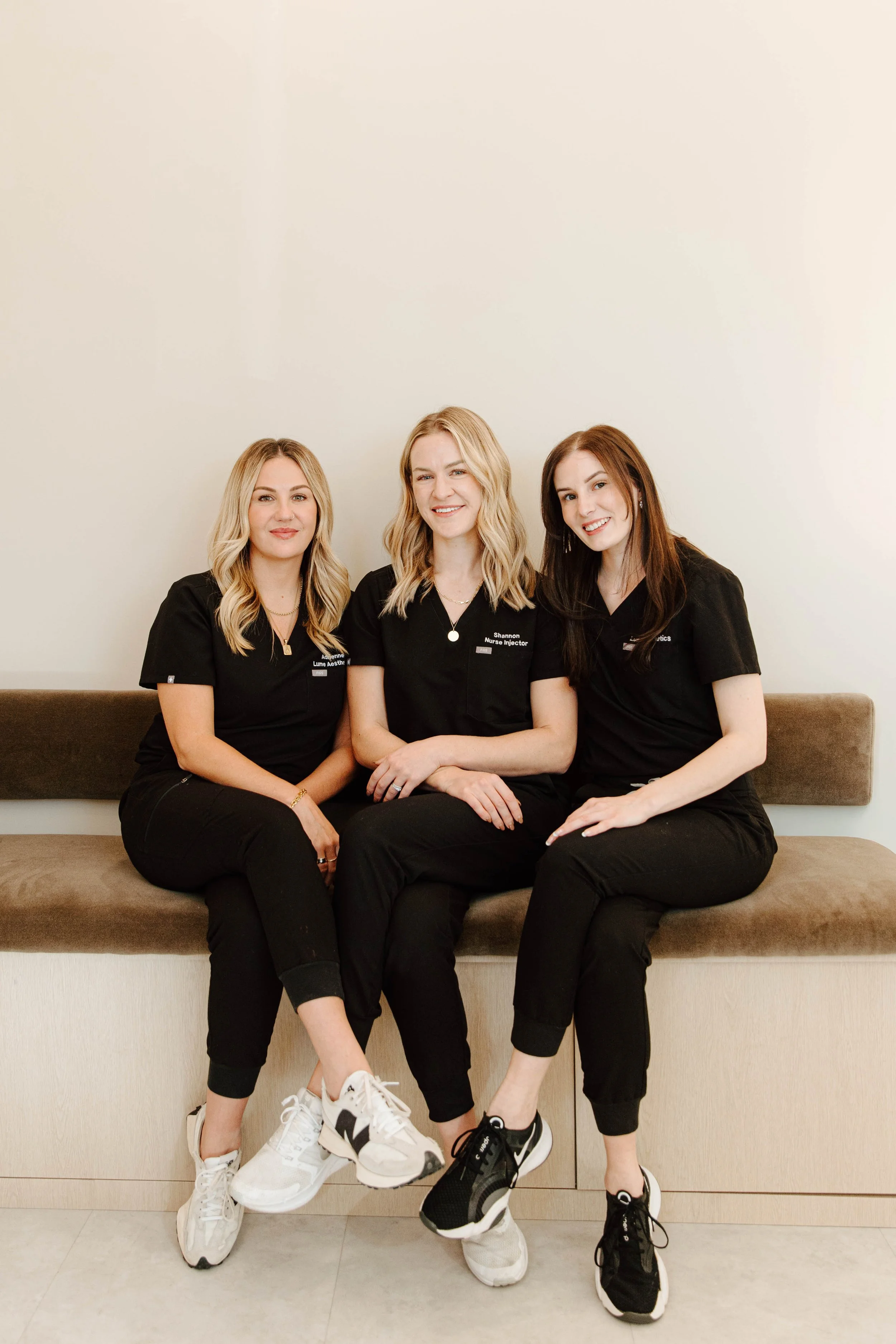 Three women in black uniforms sitting on a bench against a plain white wall.