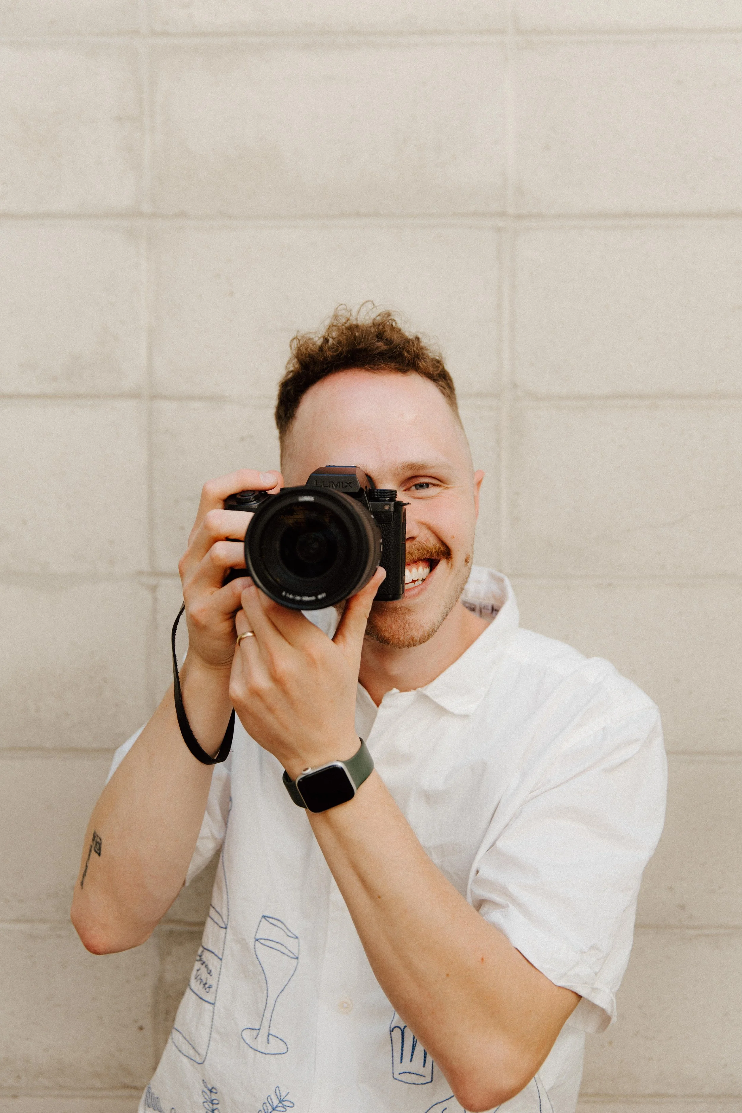 A man smiling and holding a camera up to his face, taking a picture, dressed in a white shirt with a wine and beer print, against a beige brick wall.
