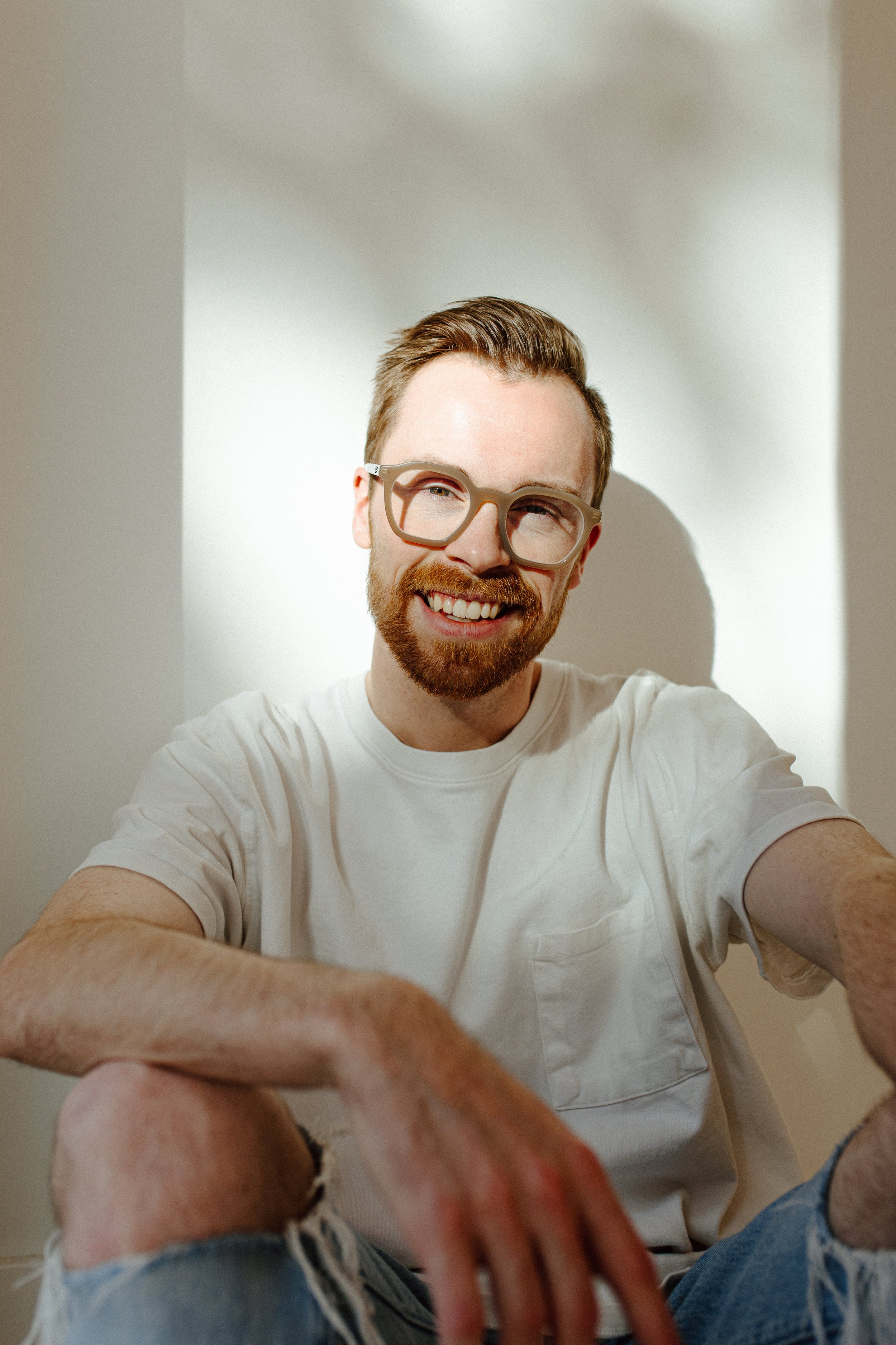 A smiling man with glasses and a beard taking a selfie, sitting with one knee up, in a casual indoor setting.