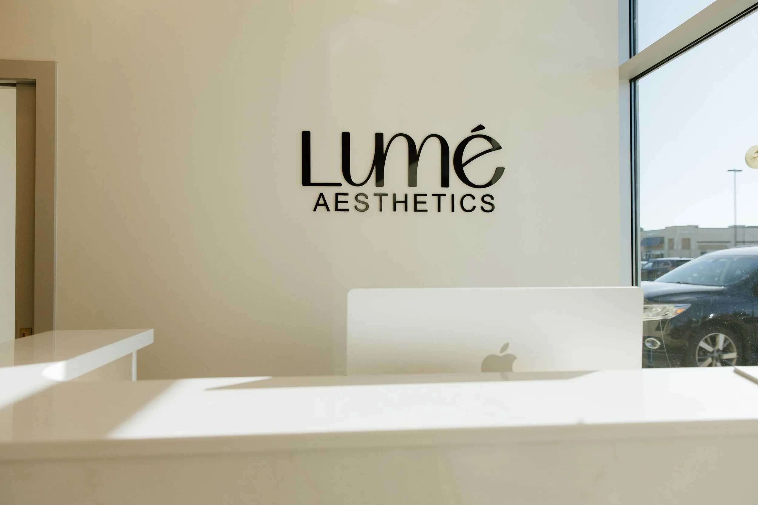 Interior of Lumé Aesthetics with a white counter, a MacBook laptop, and a wall sign displaying Lumé Aesthetics text.