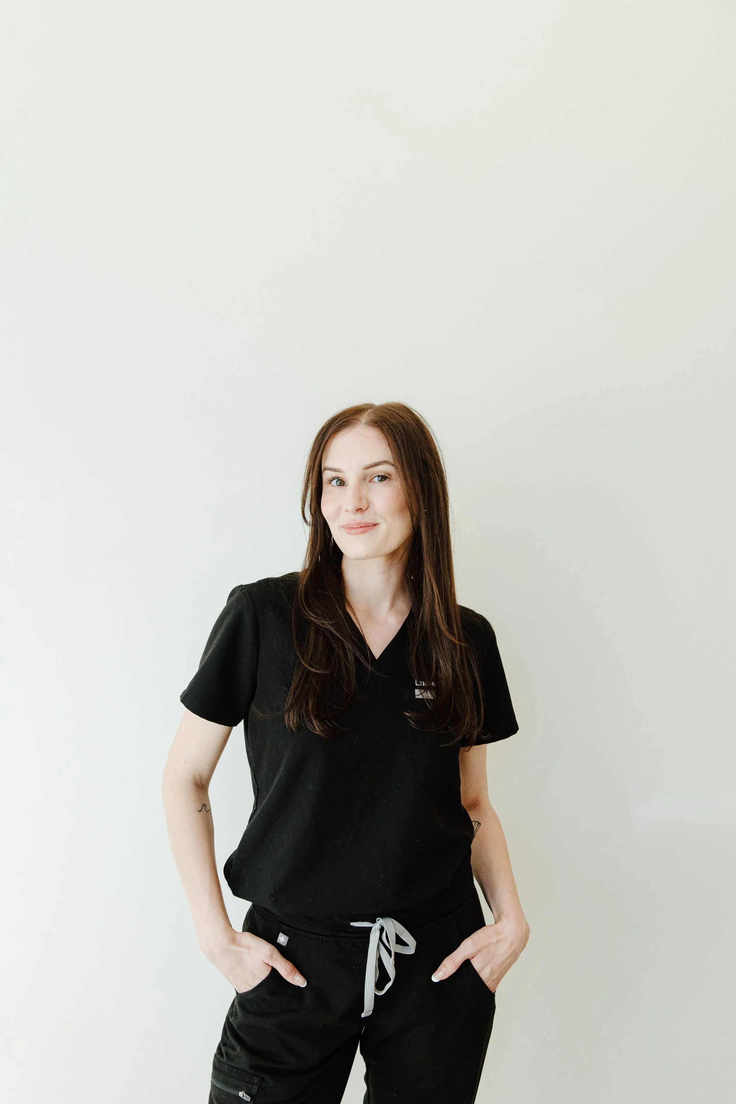 A young woman with long brown hair, wearing black medical scrubs, standing against a plain white wall, smiling slightly with hands in her pockets.