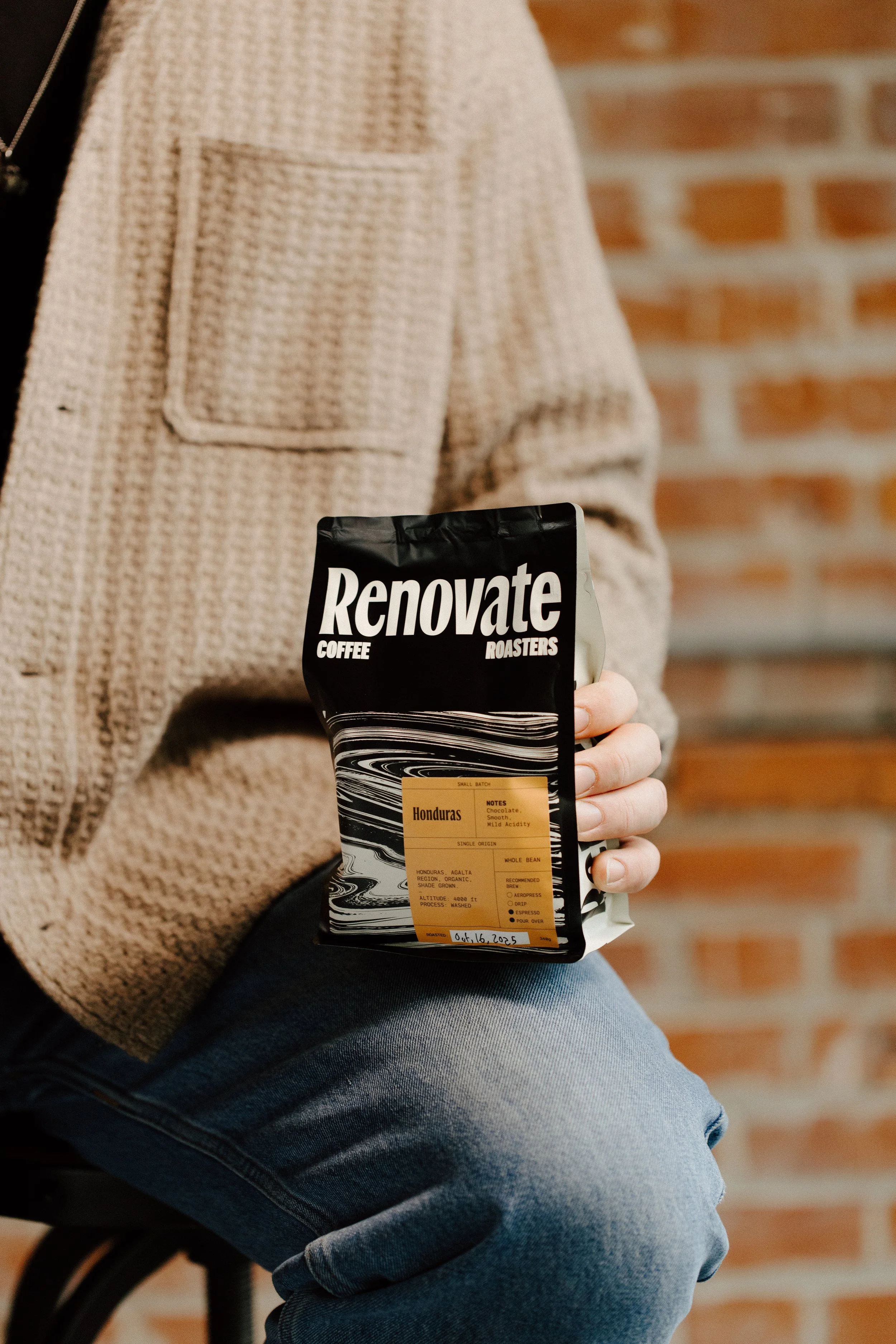 Person sitting on a stool holding a bag of Renovate Coffee Roasters Honduras single-origin coffee beans, with a brick wall in the background.