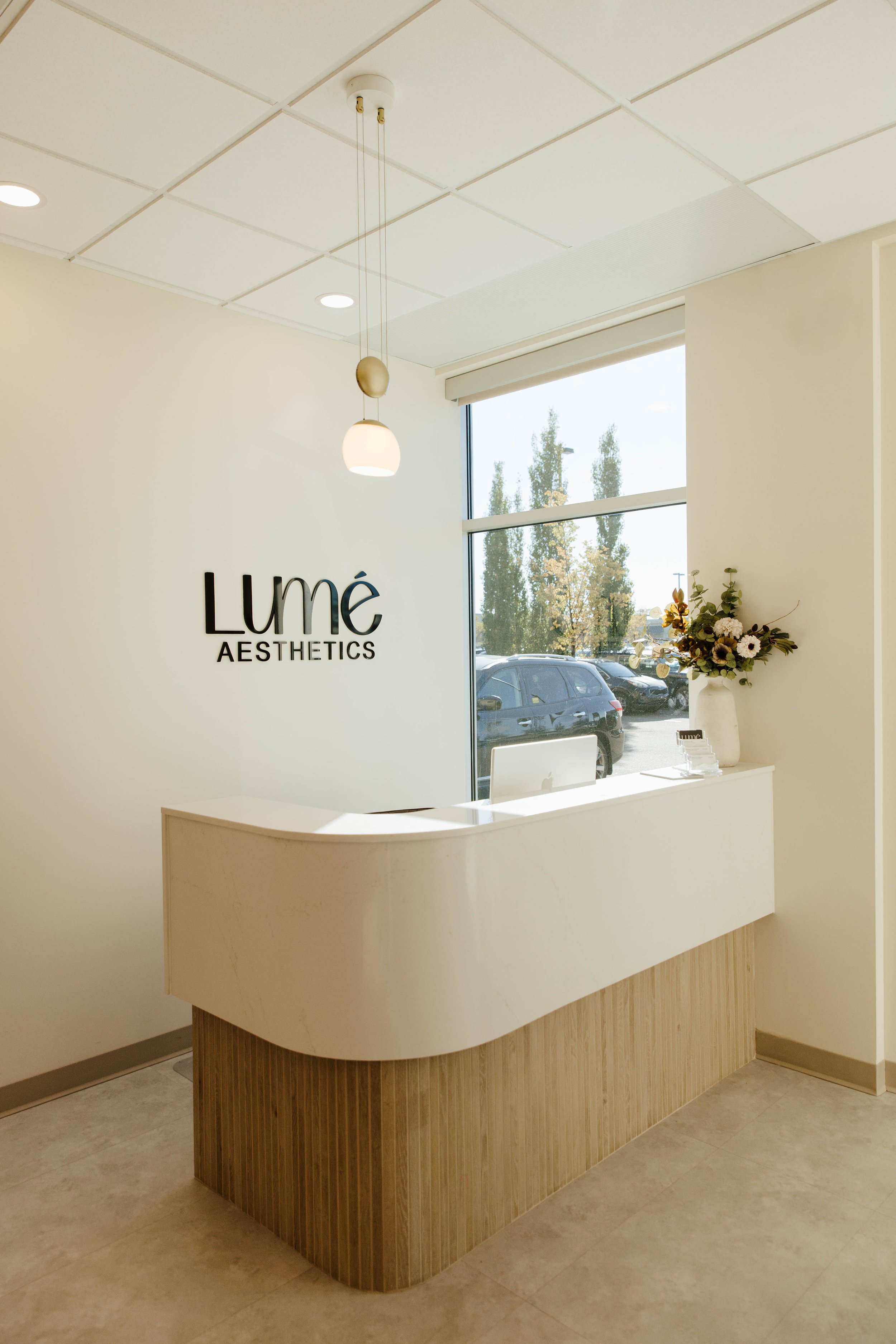 Reception desk at Lumé Aesthetics with a vase of flowers, a computer, a large window with cars outside, and a sign on the wall reading 'Lumé Aesthetics'.