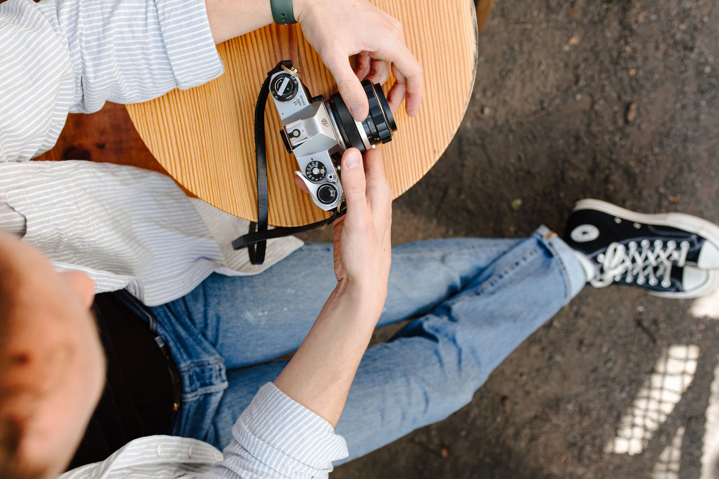 Person sitting at a round wooden table holding a vintage silver camera with a black strap, outdoors on a dirt surface.