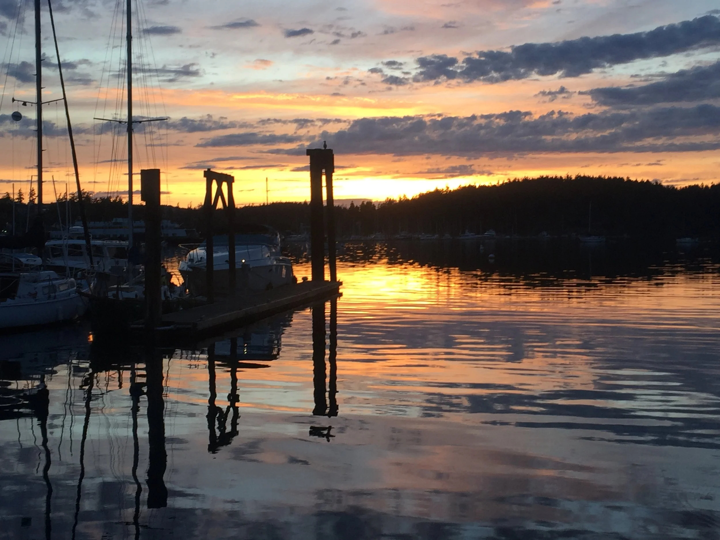 A marina at sunset with boats docked, silhouetted pilings, and a colorful sky reflected in calm water.