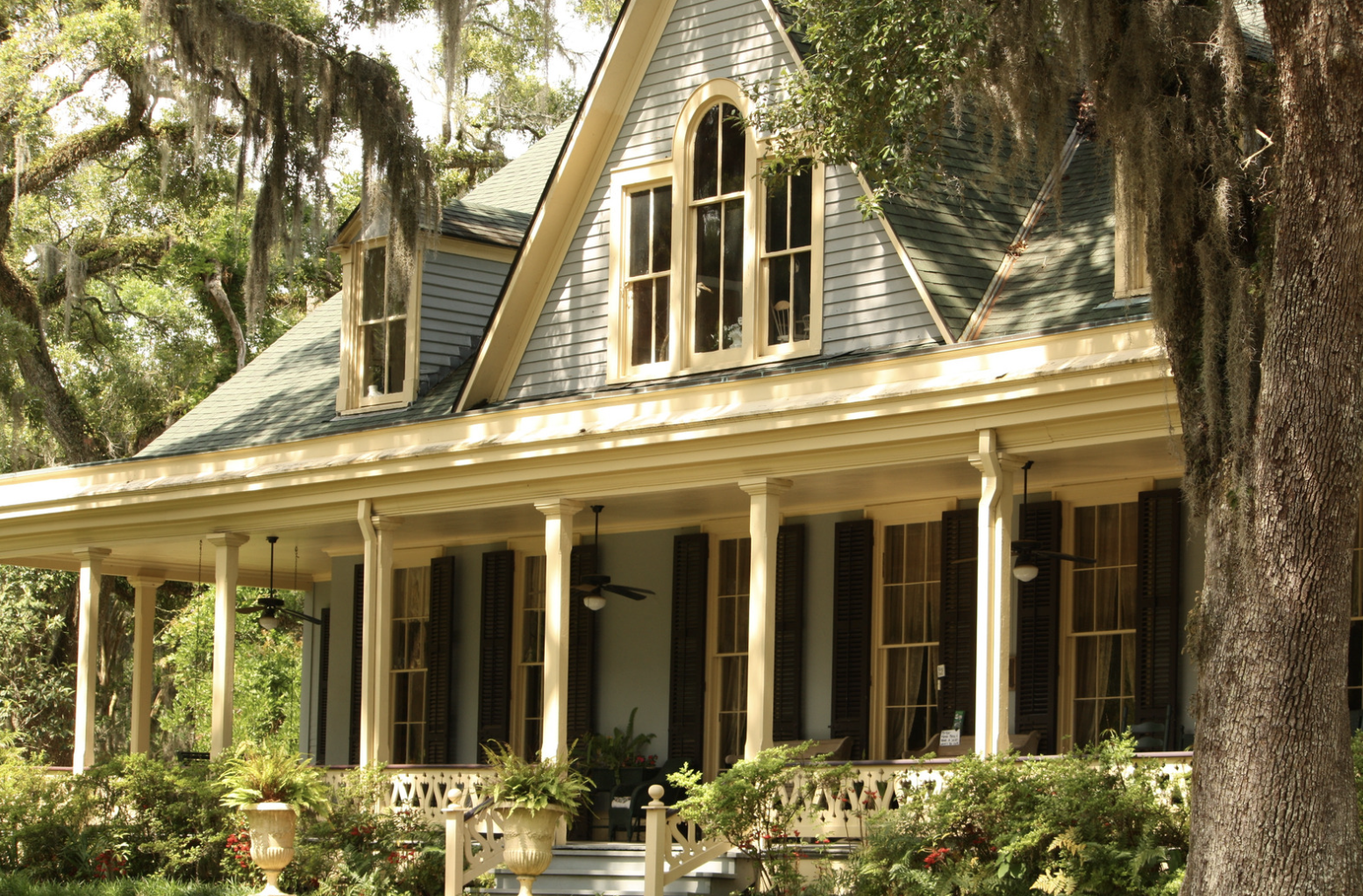 Maison ancienne à deux étages avec porche, volets en bois, fenêtres à volets en ardoise, entourée de verdure et de grands arbres.