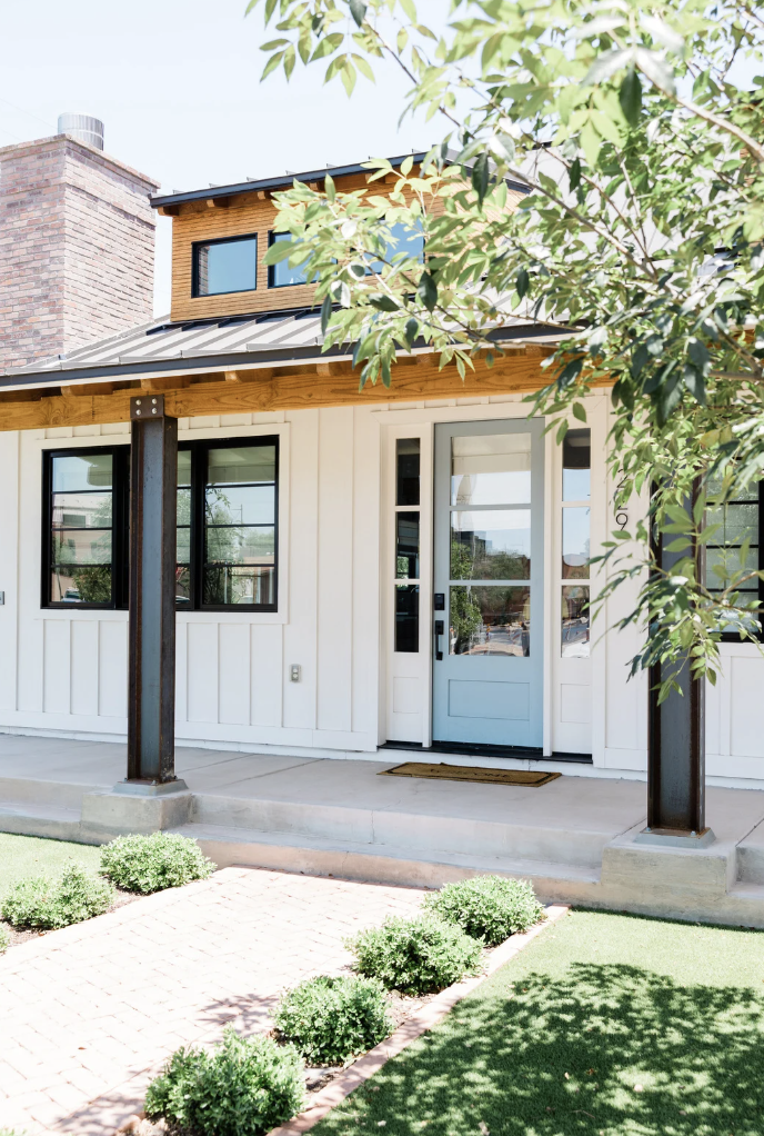 Maison moderne avec façade en bois et verres, porte d'entrée blanche, petits buissons dans la pelouse verte, arbre partiellement visible.