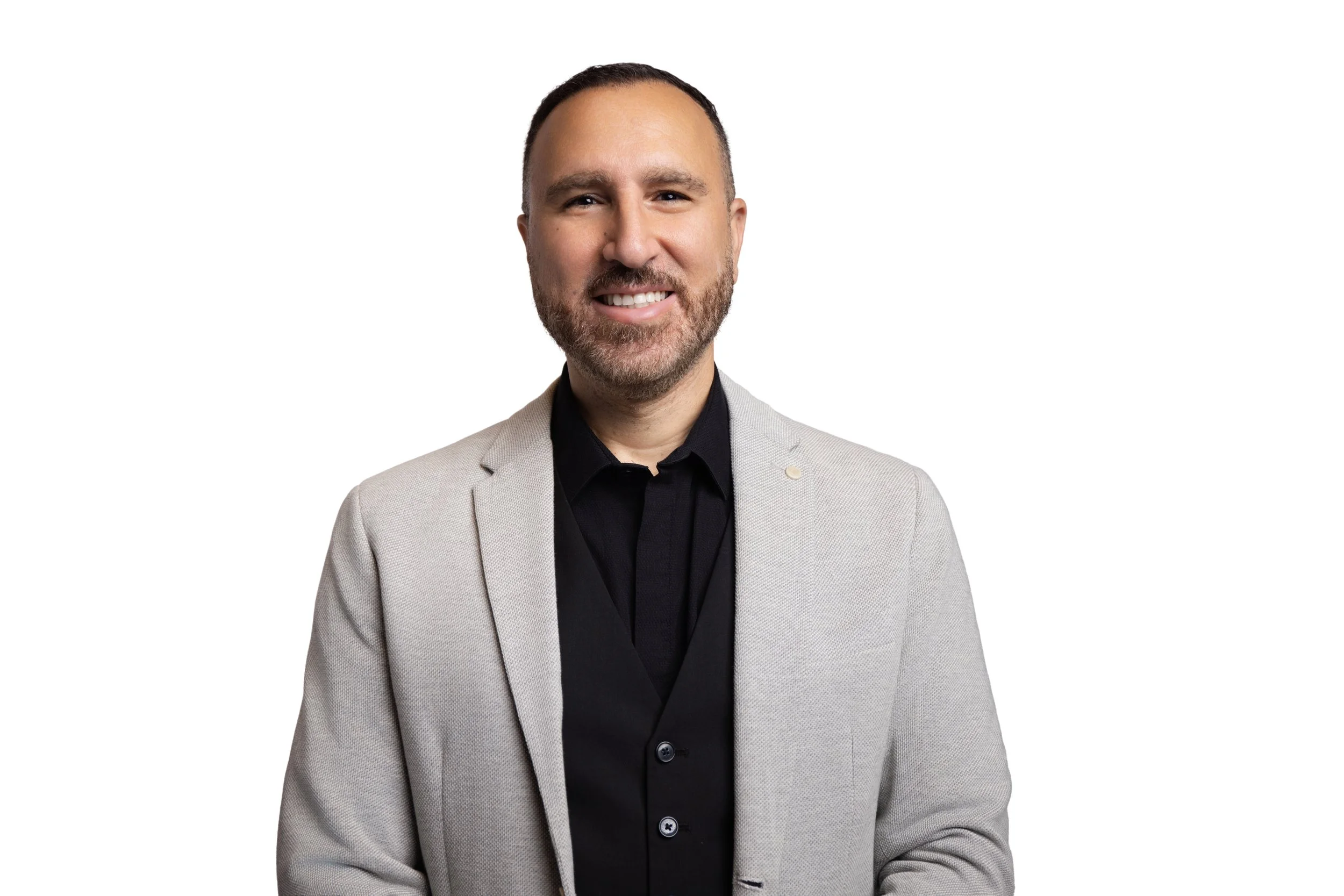 Homme d'affaires portant un costume gris et une chemise noire, souriant face à l'objectif, fond blanc.