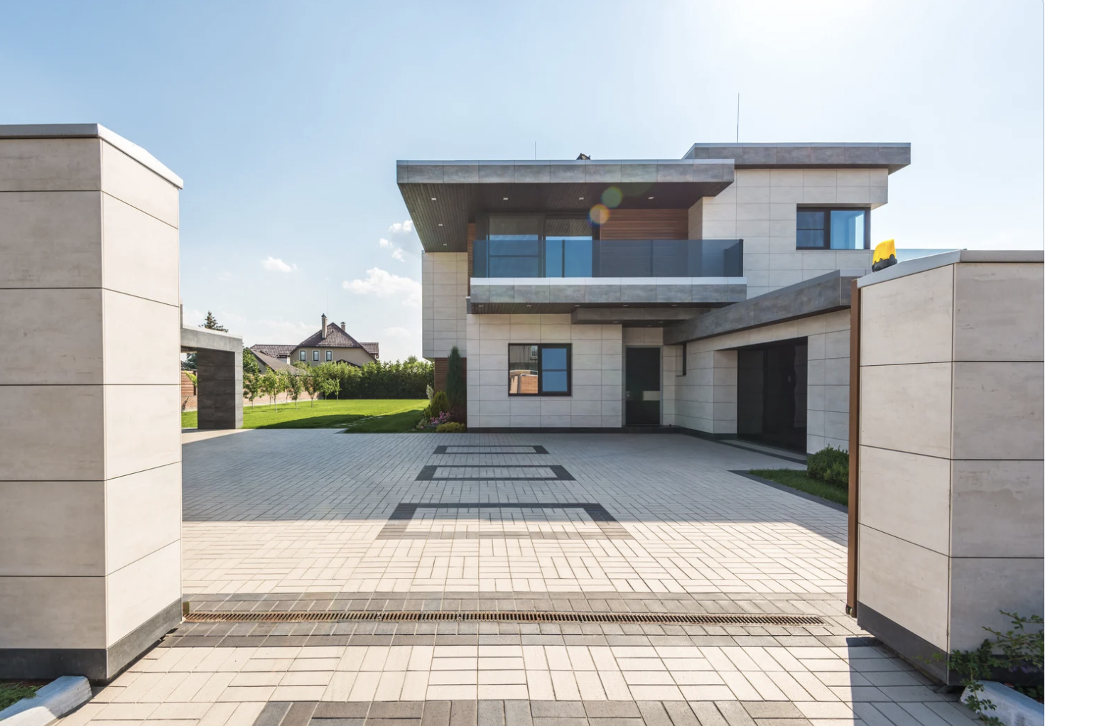 Vue de la façade moderne d'une maison avec un garage, un balcon et une allée pavée. Terrain vert et ciel en arrière-plan.