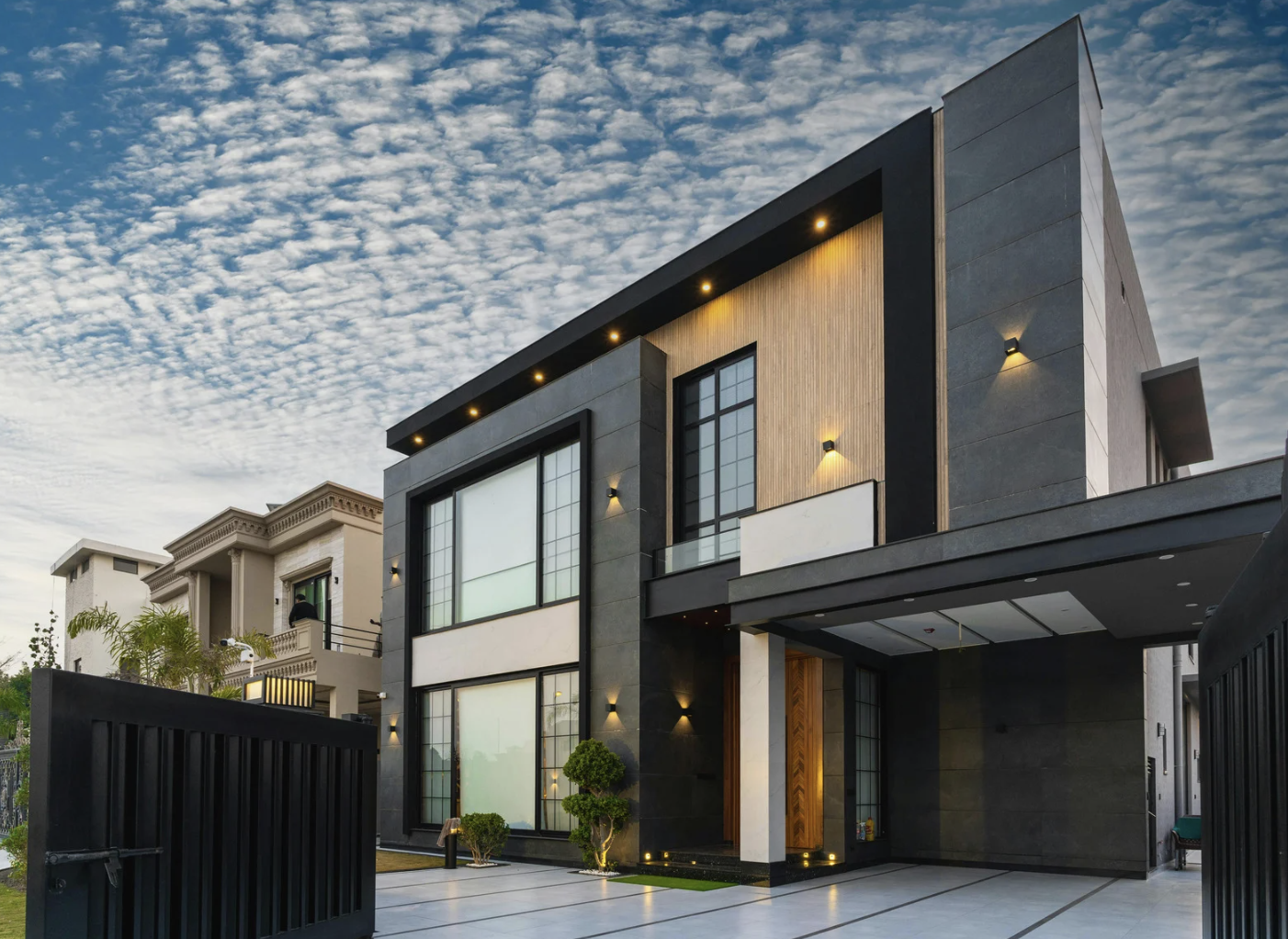 Une maison moderne à plusieurs étages avec des murs en pierre grise et en bois, des fenêtres en verre, un portail noir et un jardin bien entretenu, sous un ciel nuageux en début de soirée.
