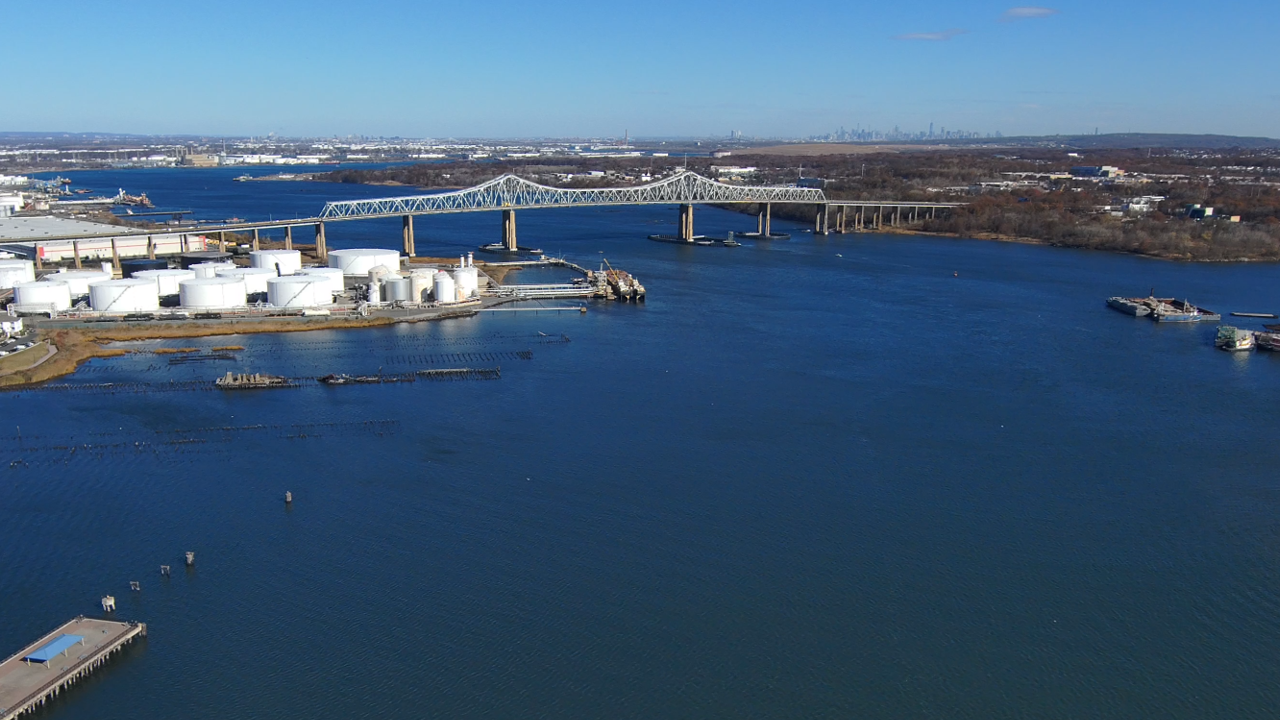 Aerial view of a river with industrial tanks, a bridge, and a city skyline in the distance.