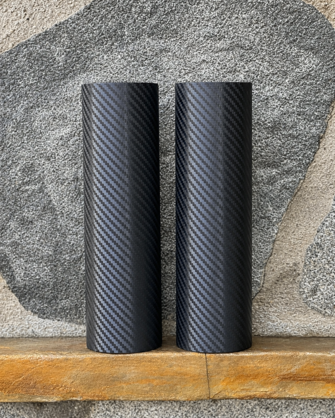 Two black RokPro motorcycle fork guards with a carbon vinyl wrap standing upright against a textured stone wall background.
