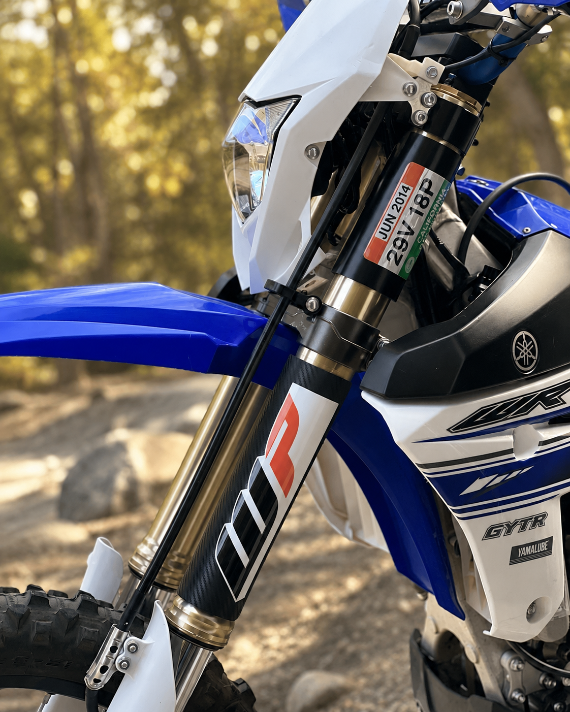 Close-up of a blue and white dirt bike displaying a set of Rokpro Fork Armor guards, outdoors on a dirt path with green trees and hills in the background.