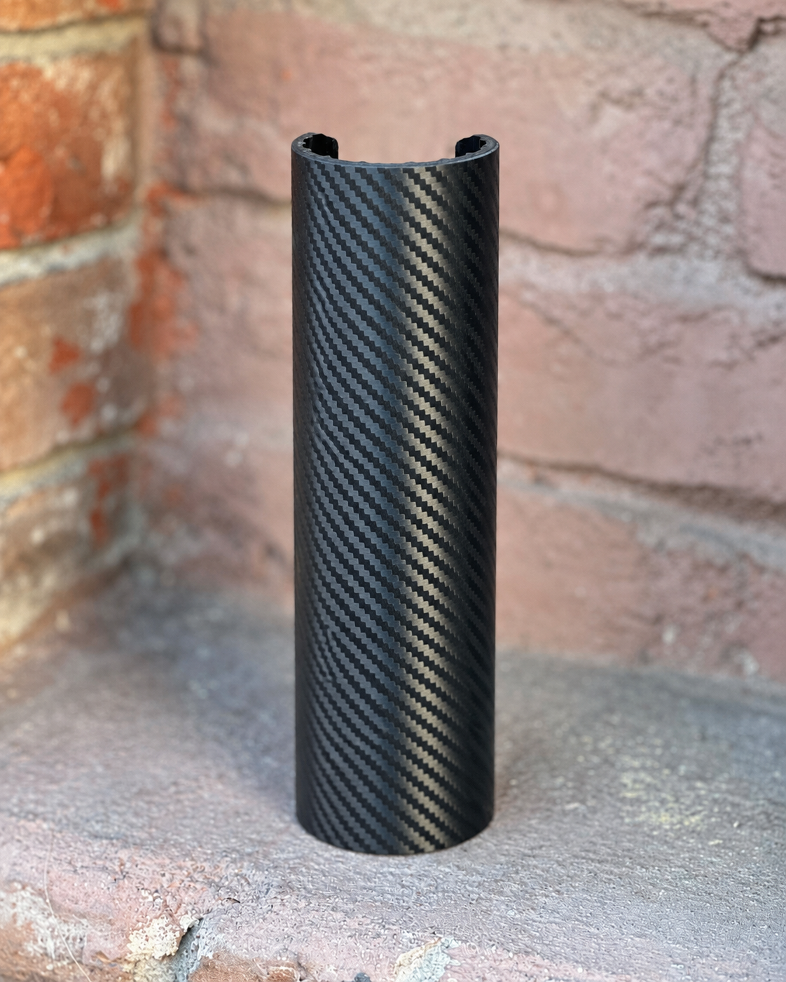 A RokPro motorcycle fork guard with a black carbon fiber armor skin, standing upright on a concrete surface against a brick wall background.