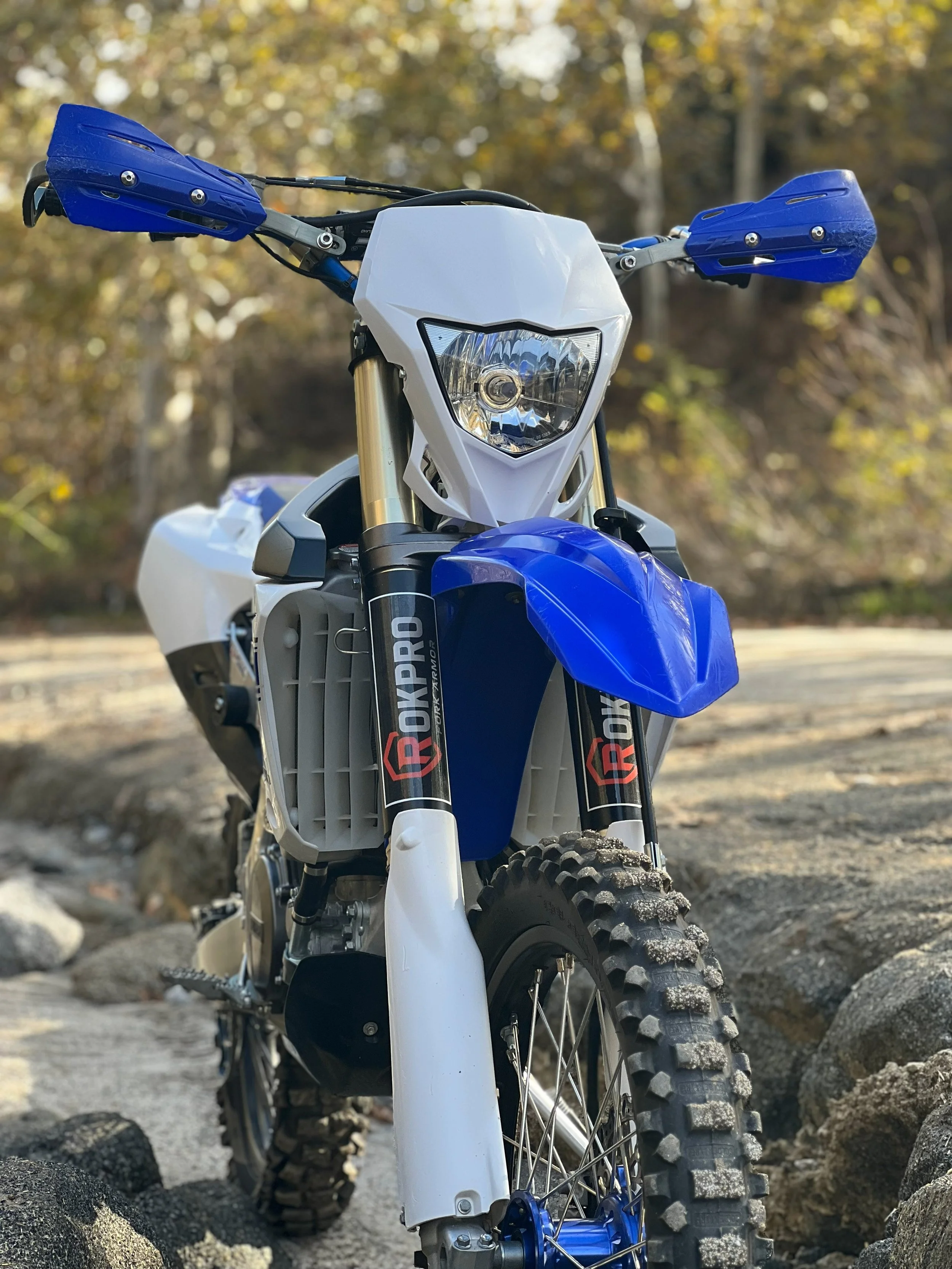 Front view of a blue and white Yamaha dirt bike motorcycle on rocky terrain with trees in the background, displaying a set of Rokpro Fork Armor guards