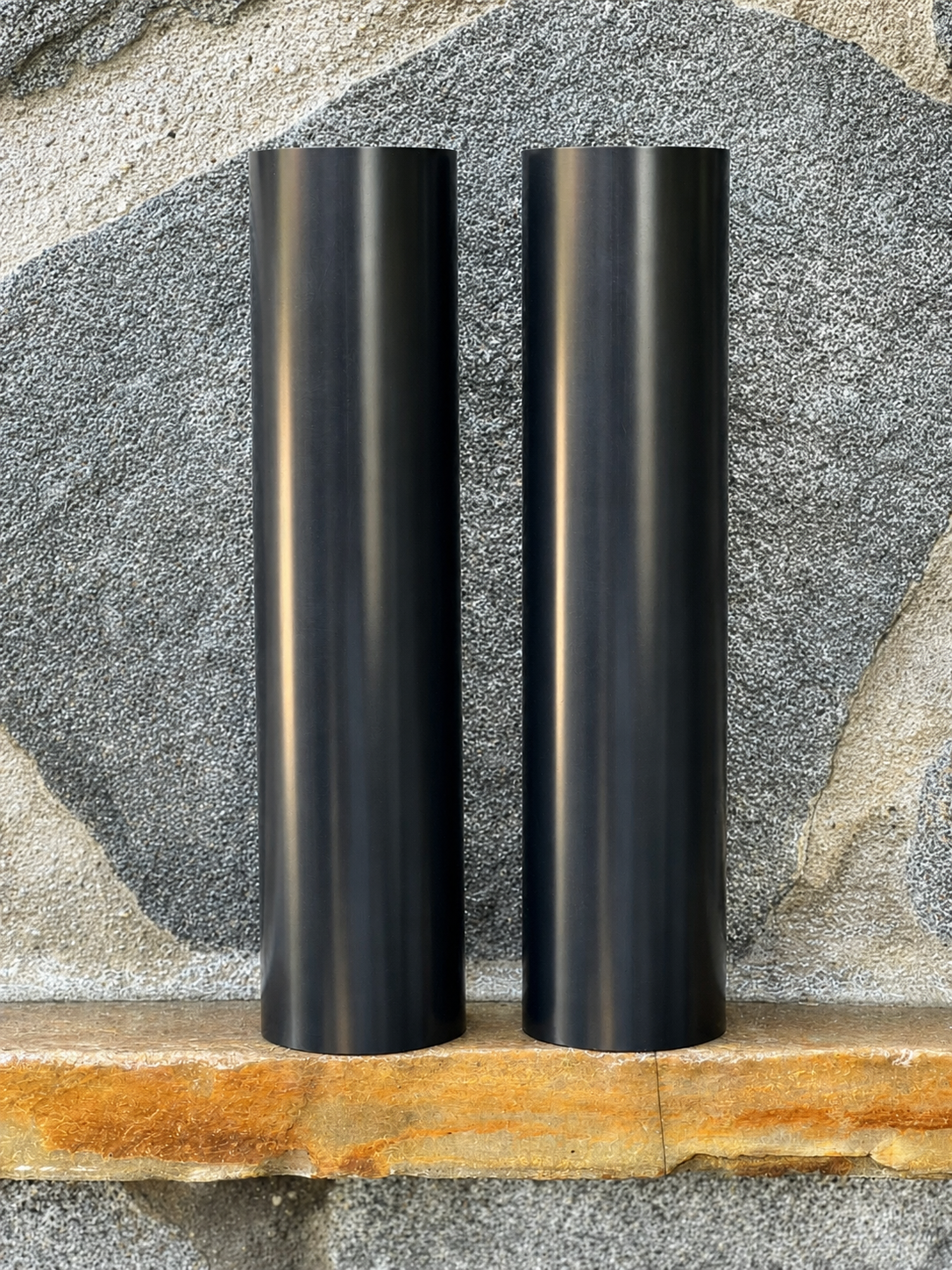 Two black RokPro motorcycle fork guards standing upright against a textured stone wall background.