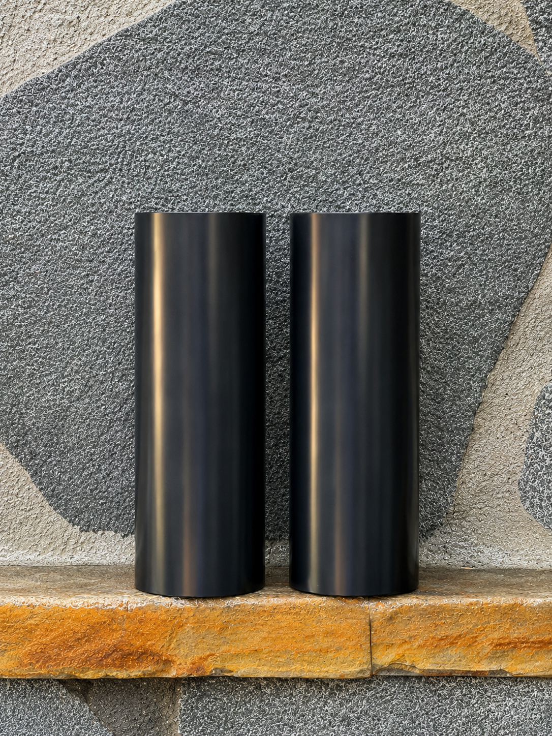 Two black RokPro motorcycle fork guards standing upright against a textured stone wall background.