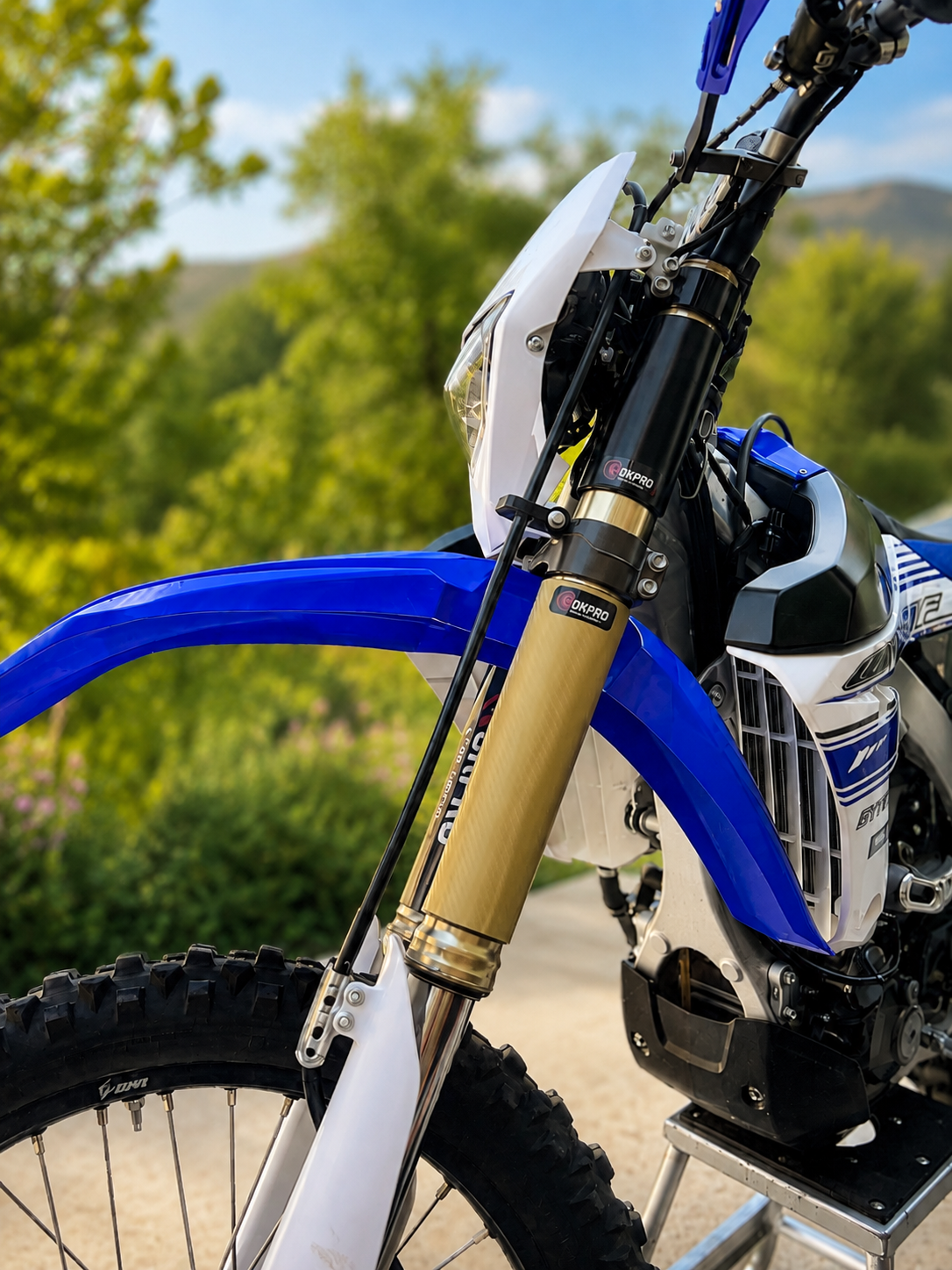Close-up of a blue and white dirt bike displaying a set of Rokpro Fork Armor guards and Armor Skins  , outdoors on a dirt path with green trees and hills in the background.