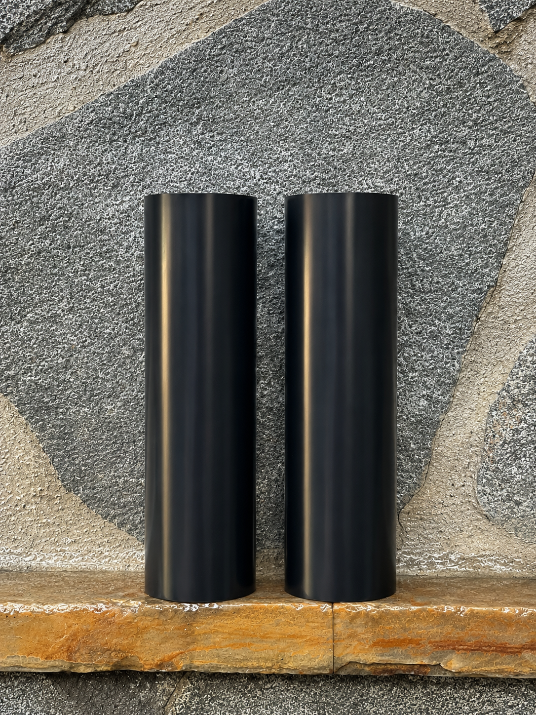 Two black RokPro motorcycle fork guards standing upright against a textured stone wall background.