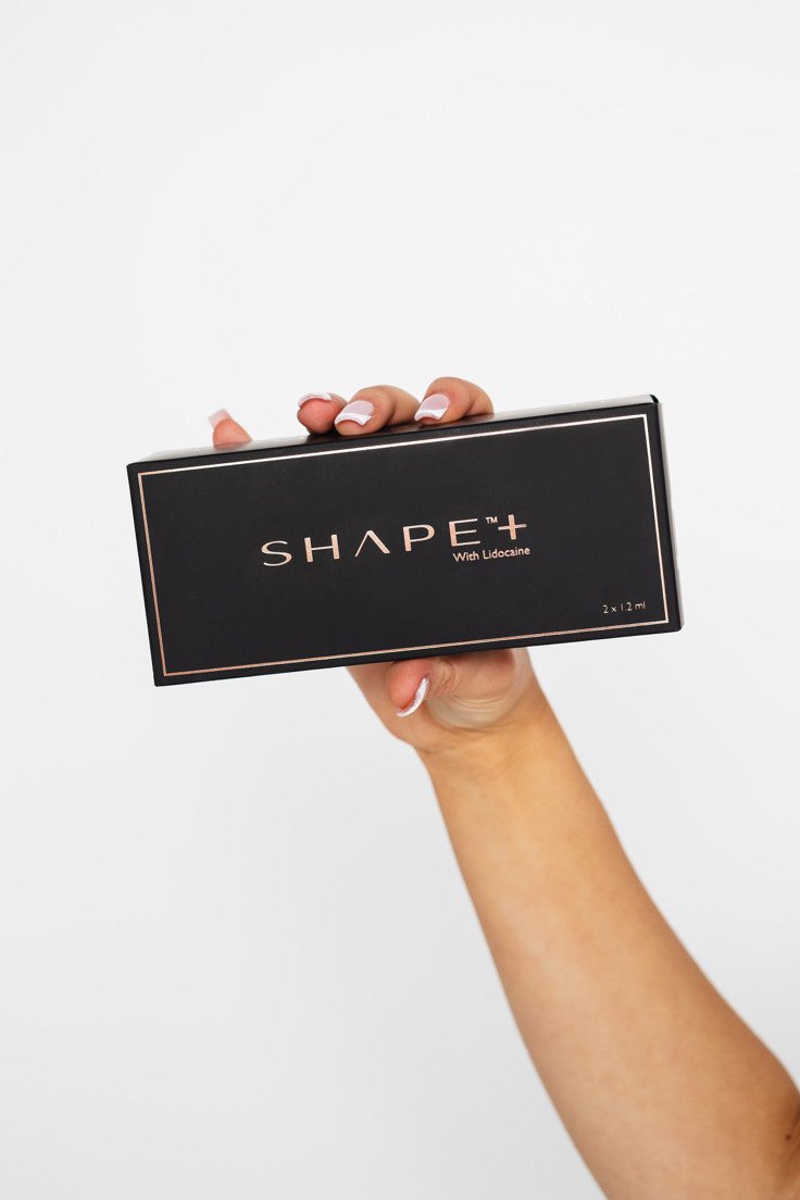A hand holding a black rectangular box labeled 'SHAPE Plus' with 'With Lidocaine' underneath, against a plain white background.