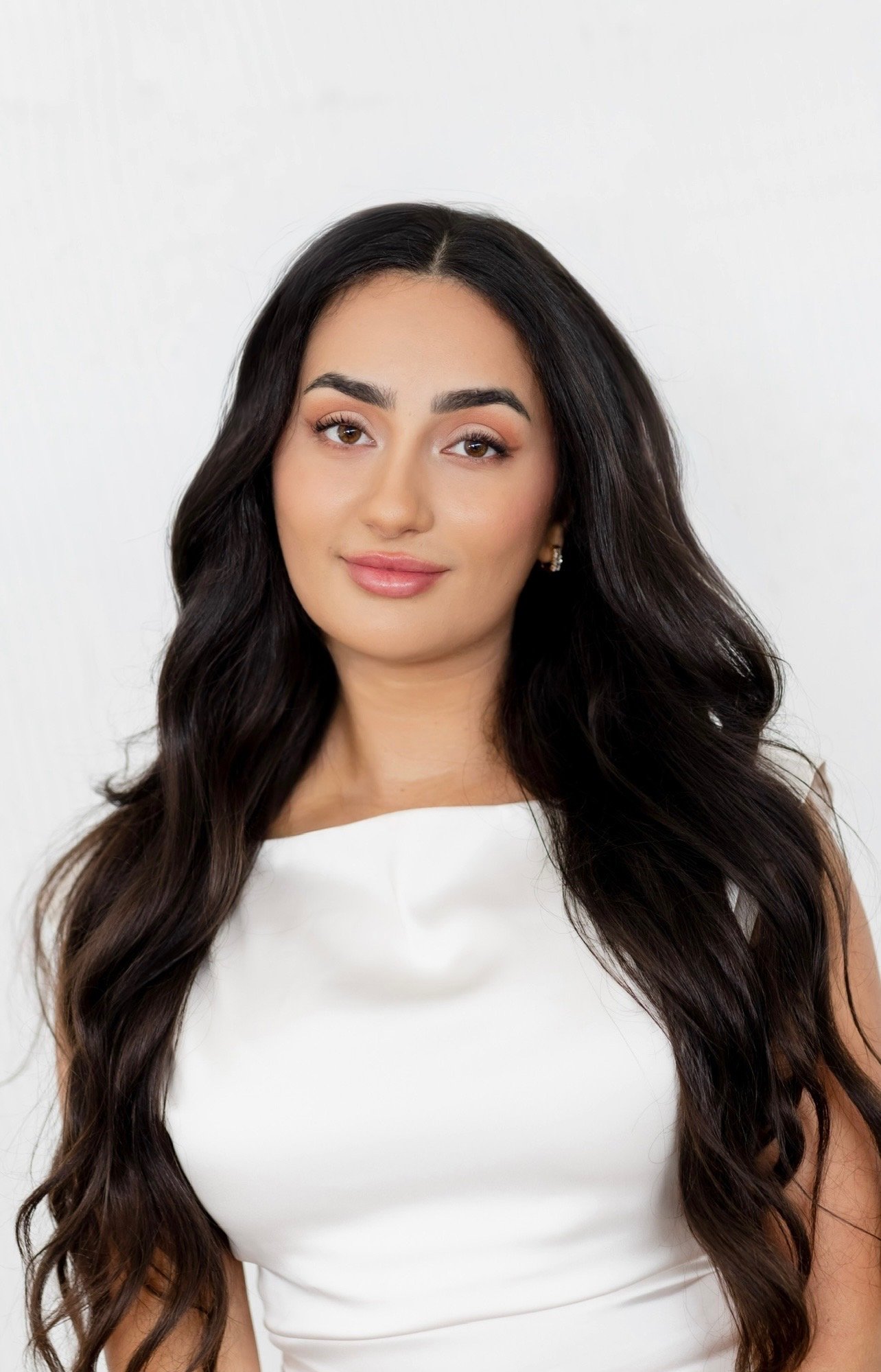 A woman with long, wavy dark hair, wearing a white sleeveless top, looking confidently at the camera.