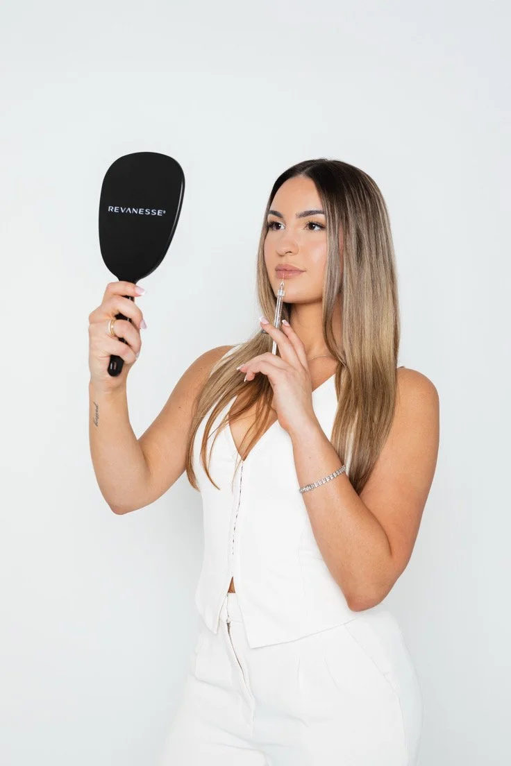 A woman with long, straight, light brown hair applying lip filler using a syringe while holding a black hand mirror with the brand name 'Revanesse'.