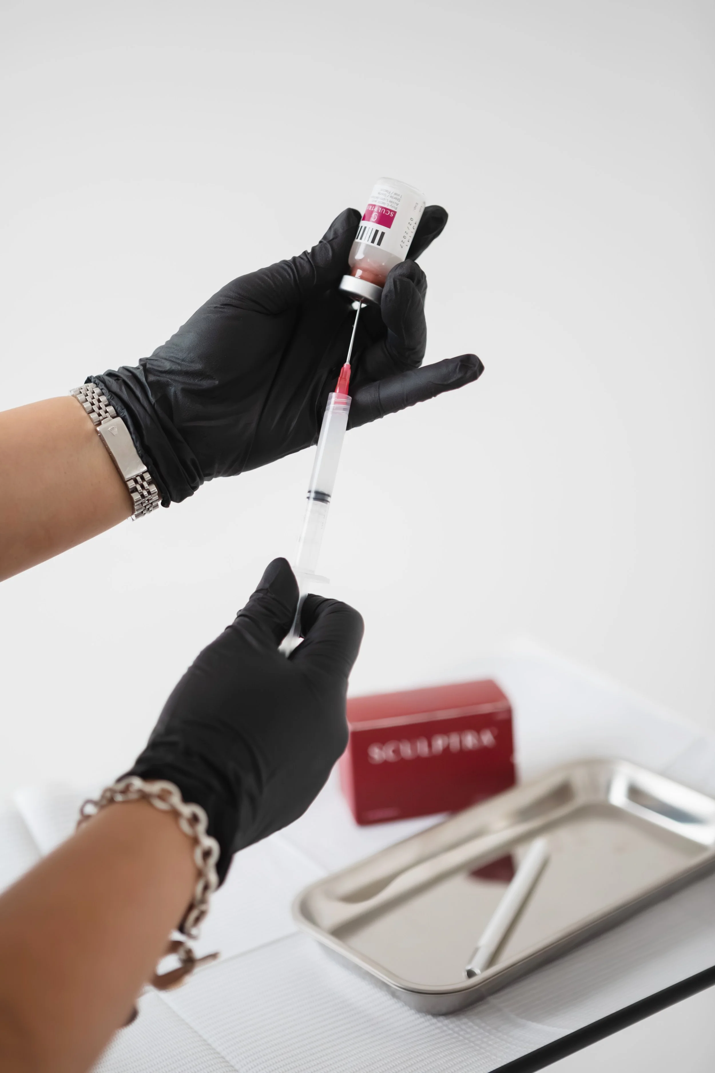 A woman receives a cosmetic facial treatment with a syringe, with a box and vial of Sculptra on the table beside her.